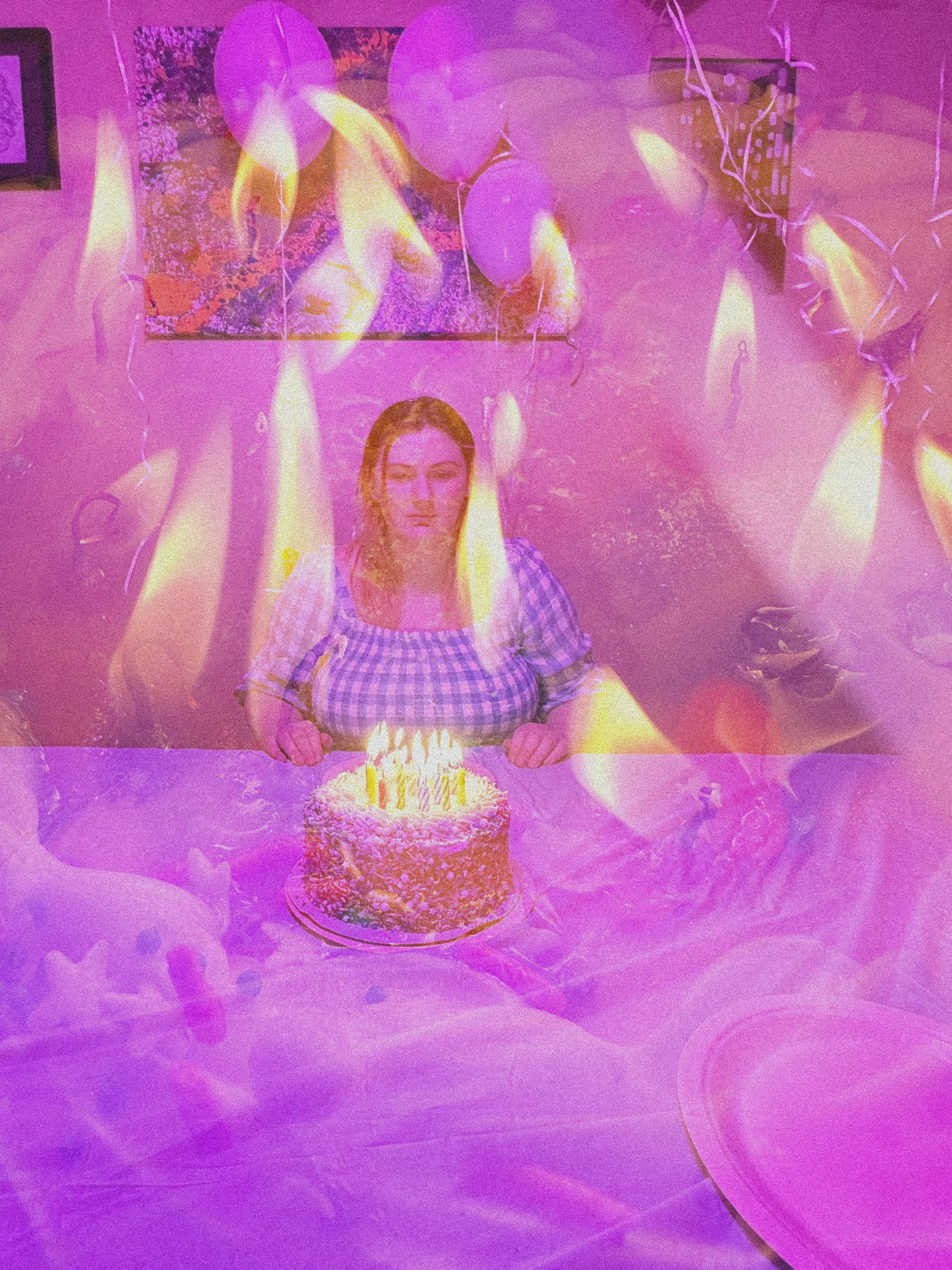 A woman sitting at a table with a birthday cake and lit candles, surrounded by purple balloons, with a colorful abstract painting on the wall behind her.