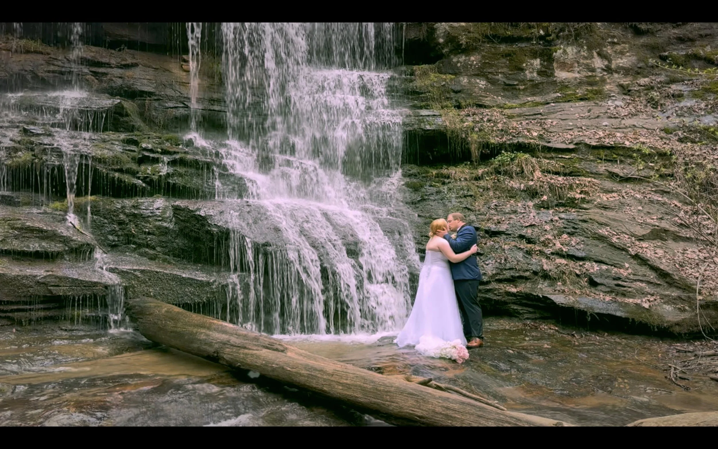 A bride and groom hugging and kissing in front of a waterfall at a scenic outdoor location, with a log on the rocks in the stream.