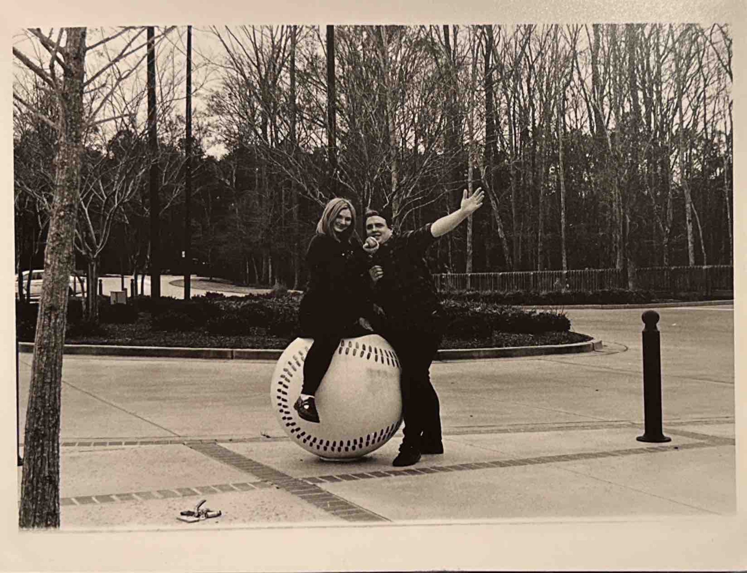 Two people sit together on a large spherical sculpture outside, with trees and a paved walkway in the background.