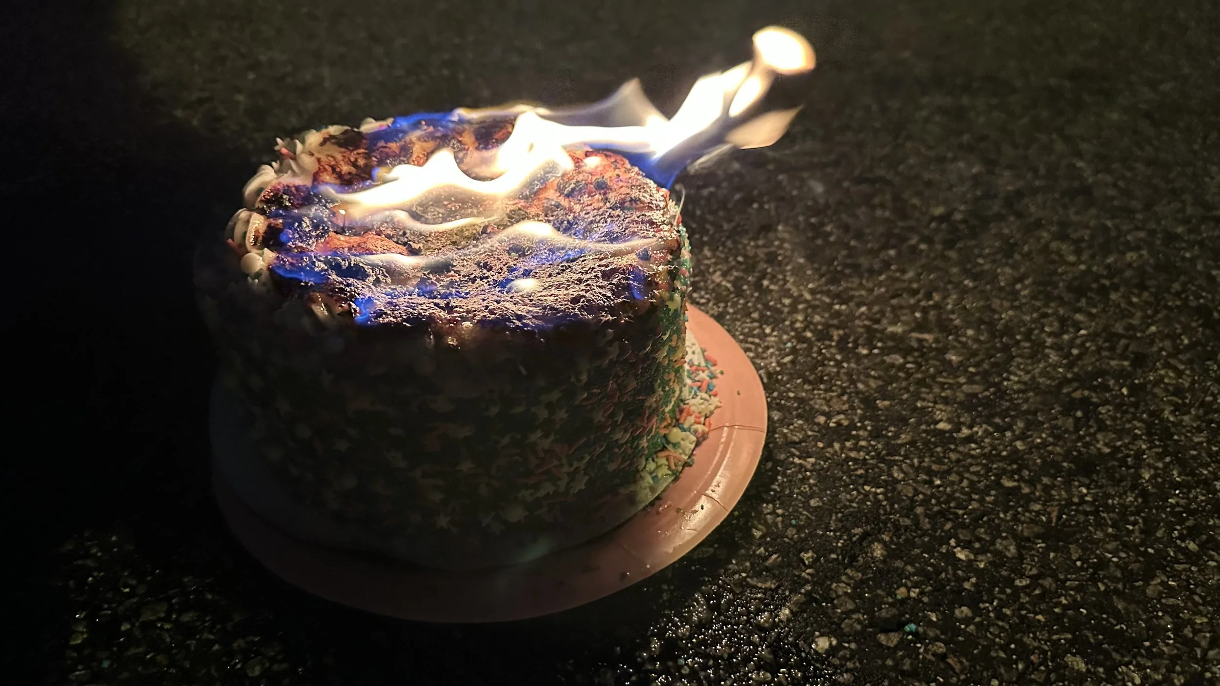 A birthday cake with lit matches on top, burning with what appears to be flames. The cake is decorated with colorful sprinkles and is placed on a round pink base on a dark surface.