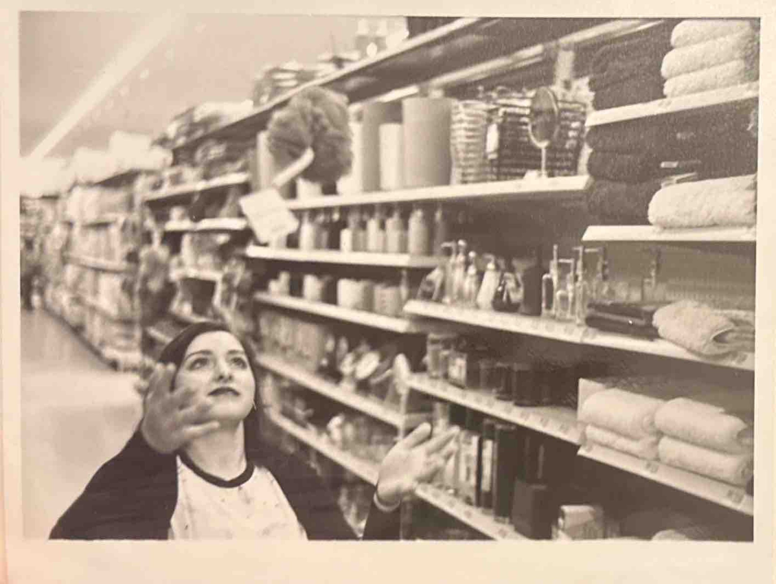 A woman with long dark hair reaching up with one hand in a supermarket aisle filled with shelves of various household items and towels.