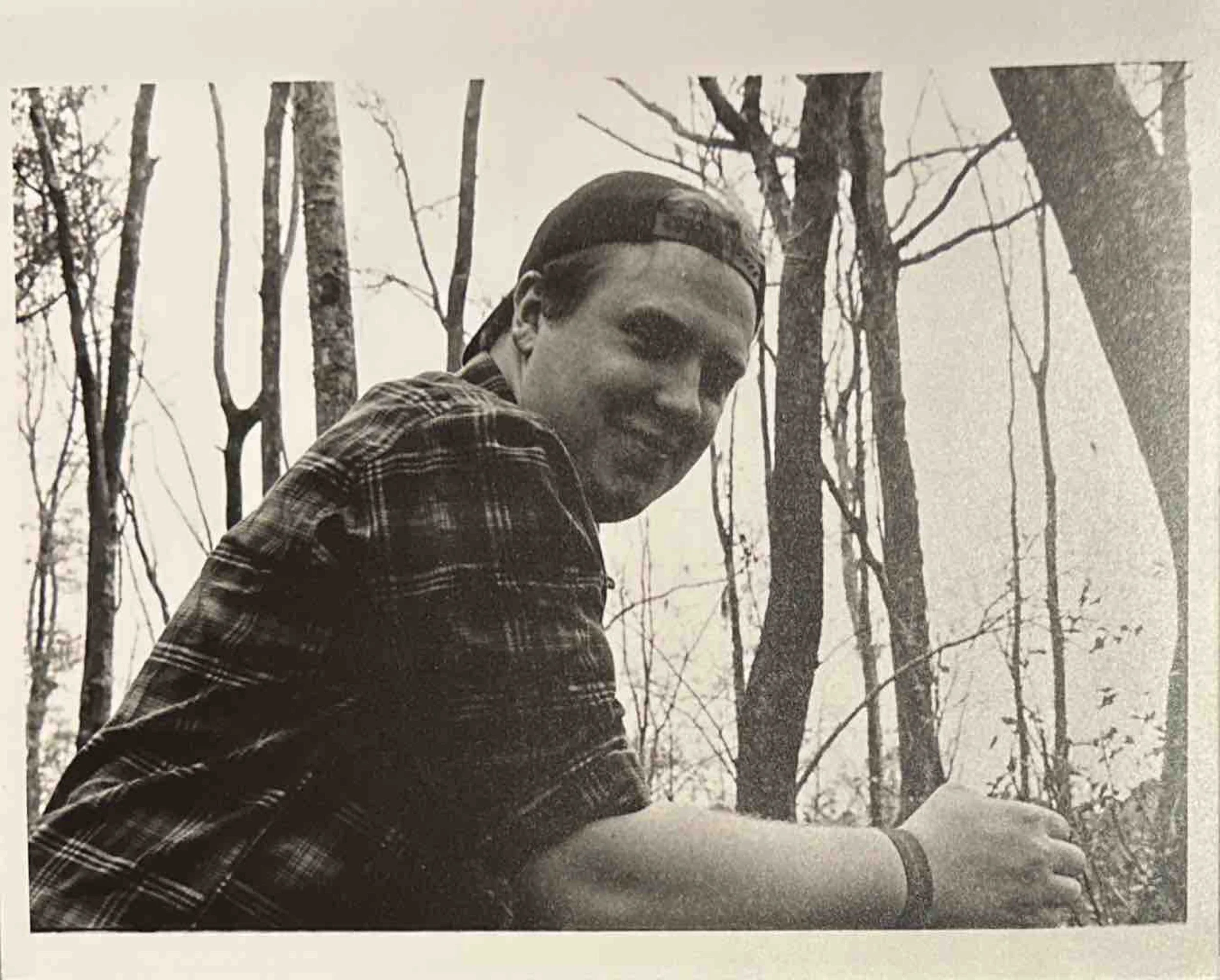 Black and white photograph of a young man wearing a baseball cap backwards and a plaid shirt, sitting outdoors among leafless trees, smiling at the camera.