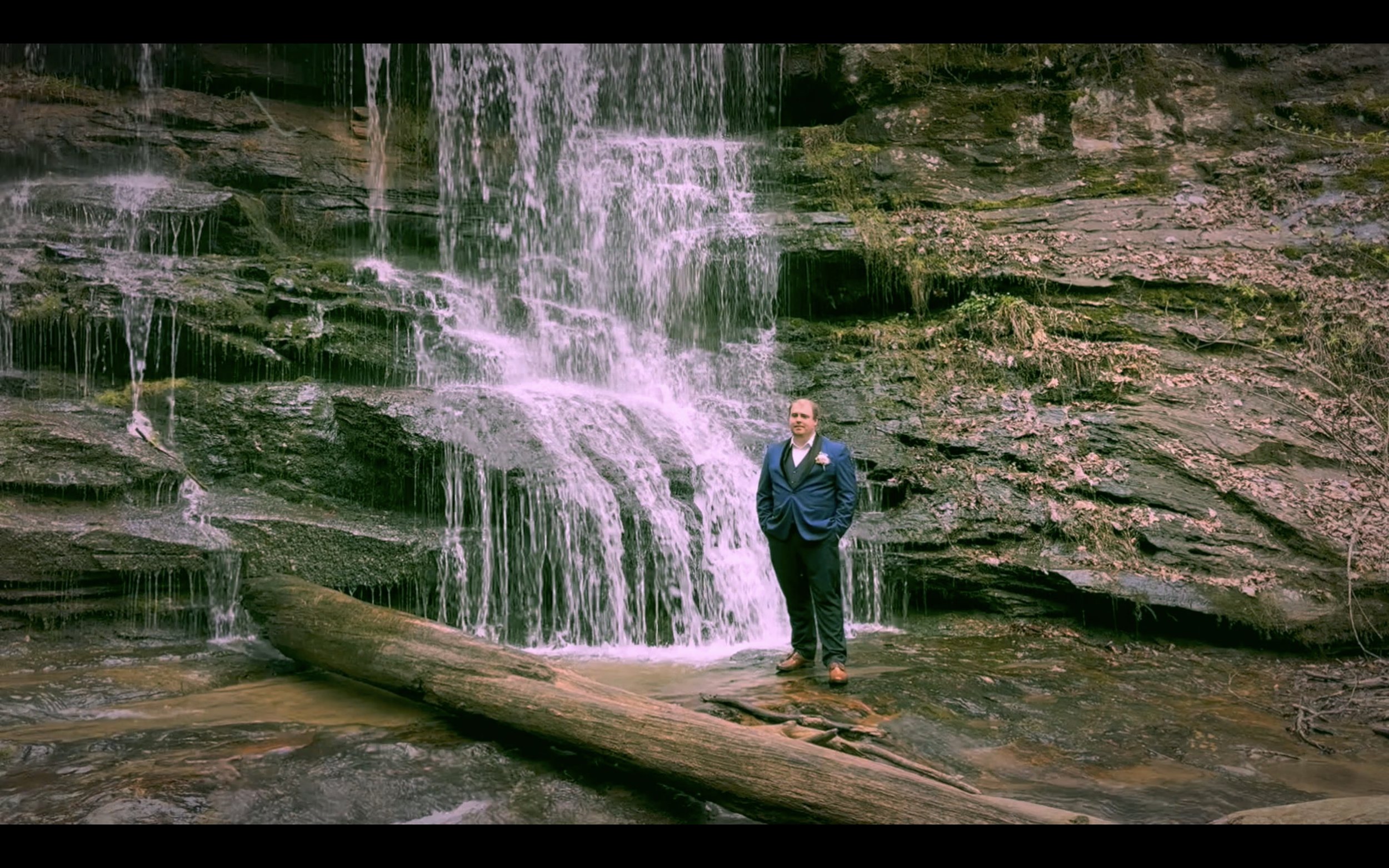 A man in a blue suit and tie standing on a large log in front of a small waterfall in a natural outdoor setting.