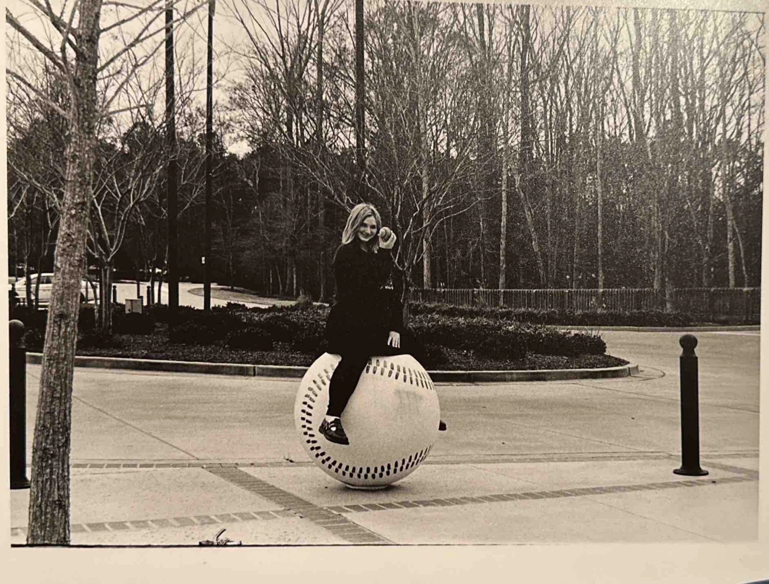 A woman sitting on a large spherical object outdoors, holding a phone to her ear and smiling, with trees and a sidewalk in the background.