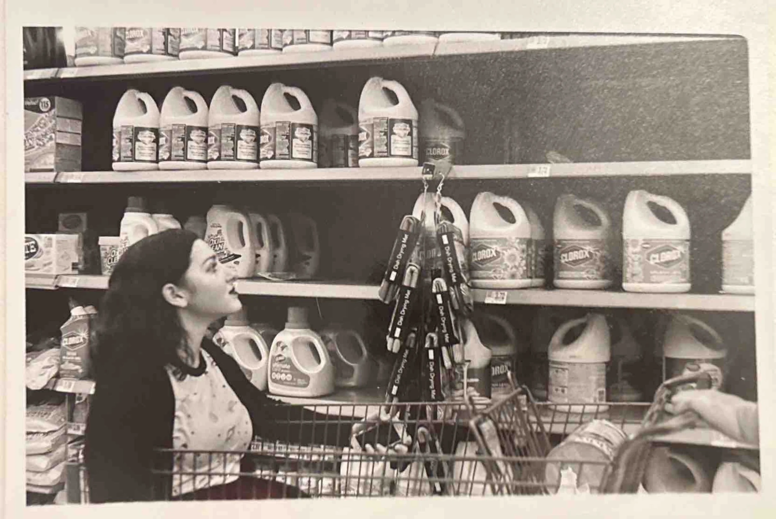 A woman in a shopping cart at a store aisle, looking up at shelves filled with cleaning products and supplies, including bottles and containers of bleach and other household chemicals.