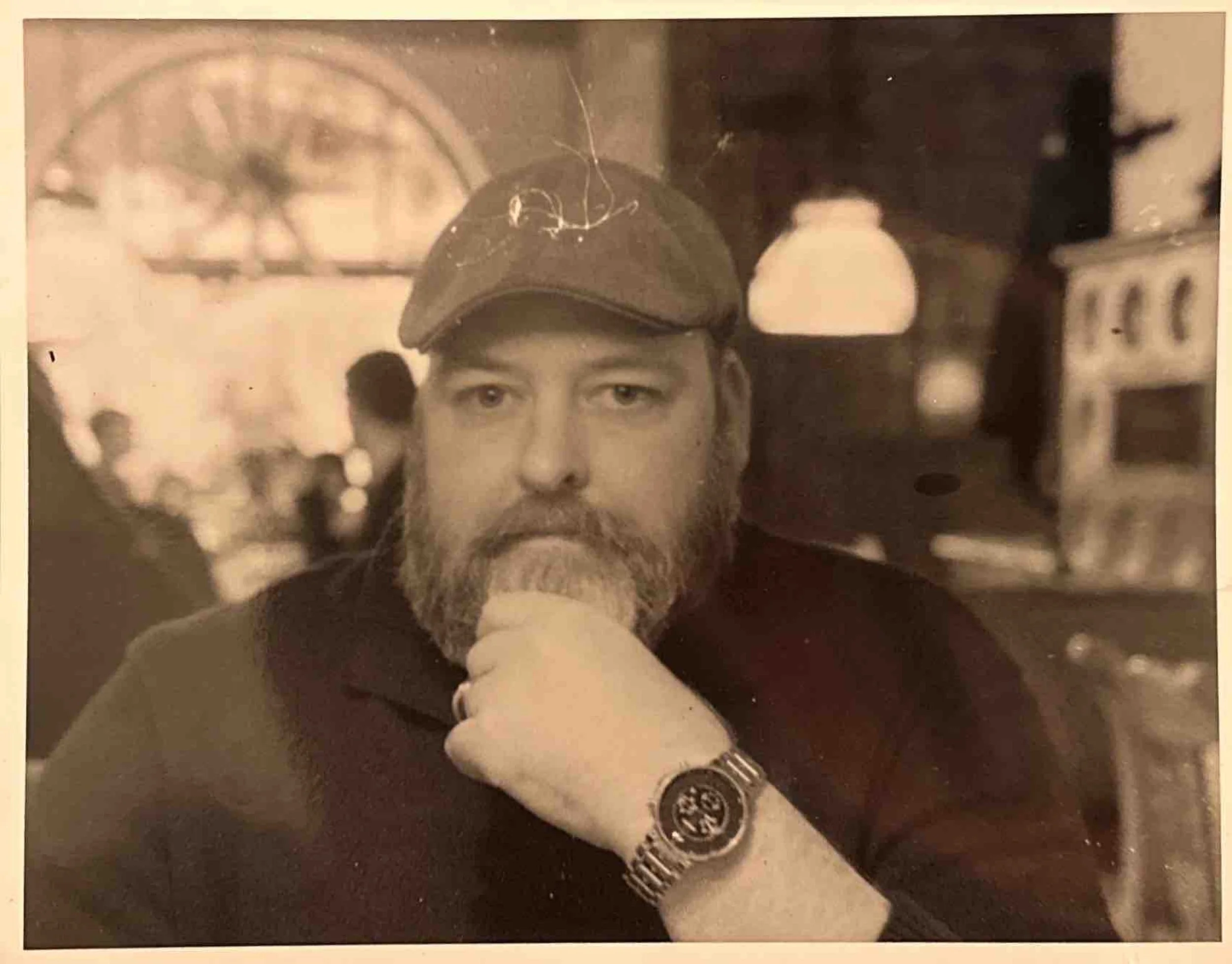 A black-and-white photograph of a light-skinned man with a beard, wearing a cap and a watch, sitting indoors with his hand on his chin, looking at the camera.