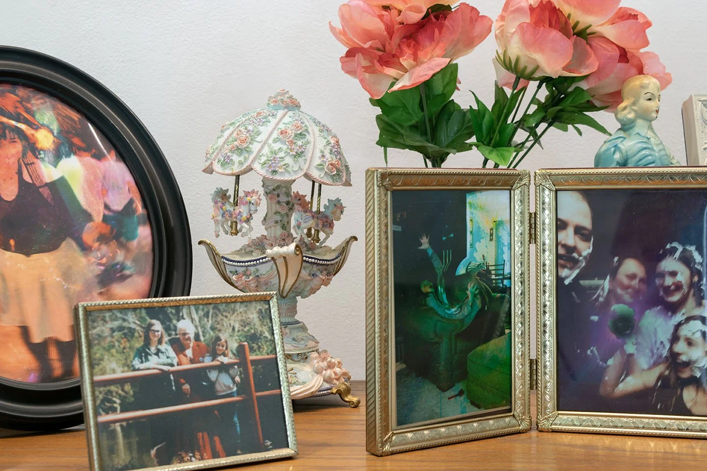 Display of framed photographs, a vintage-style ceramic carousel lamp, artificial pink flowers in a golden frame, and porcelain figurines on a wooden surface.