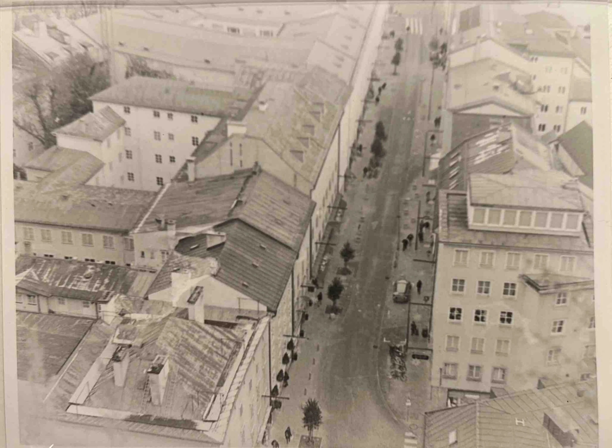 A black-and-white aerial view of a city street, showing buildings, trees, parked cars, and people walking.