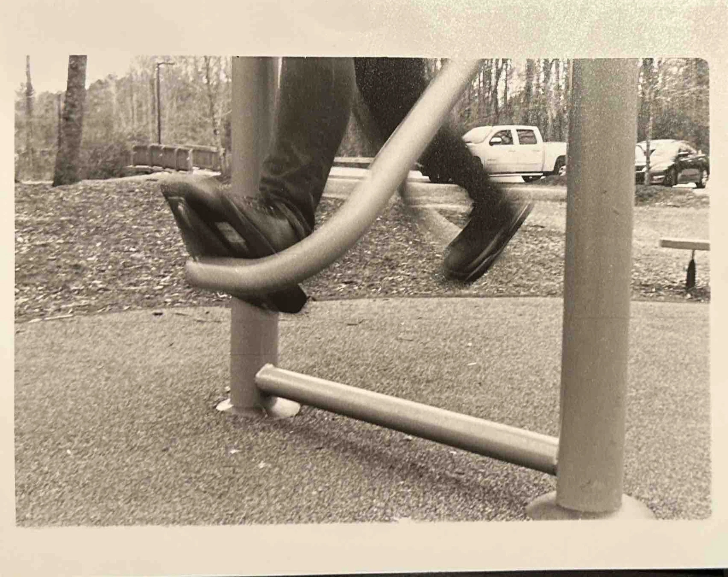 Child wearing black boots and jeans, riding a swing in a park. Park has trees and parked cars in the background.