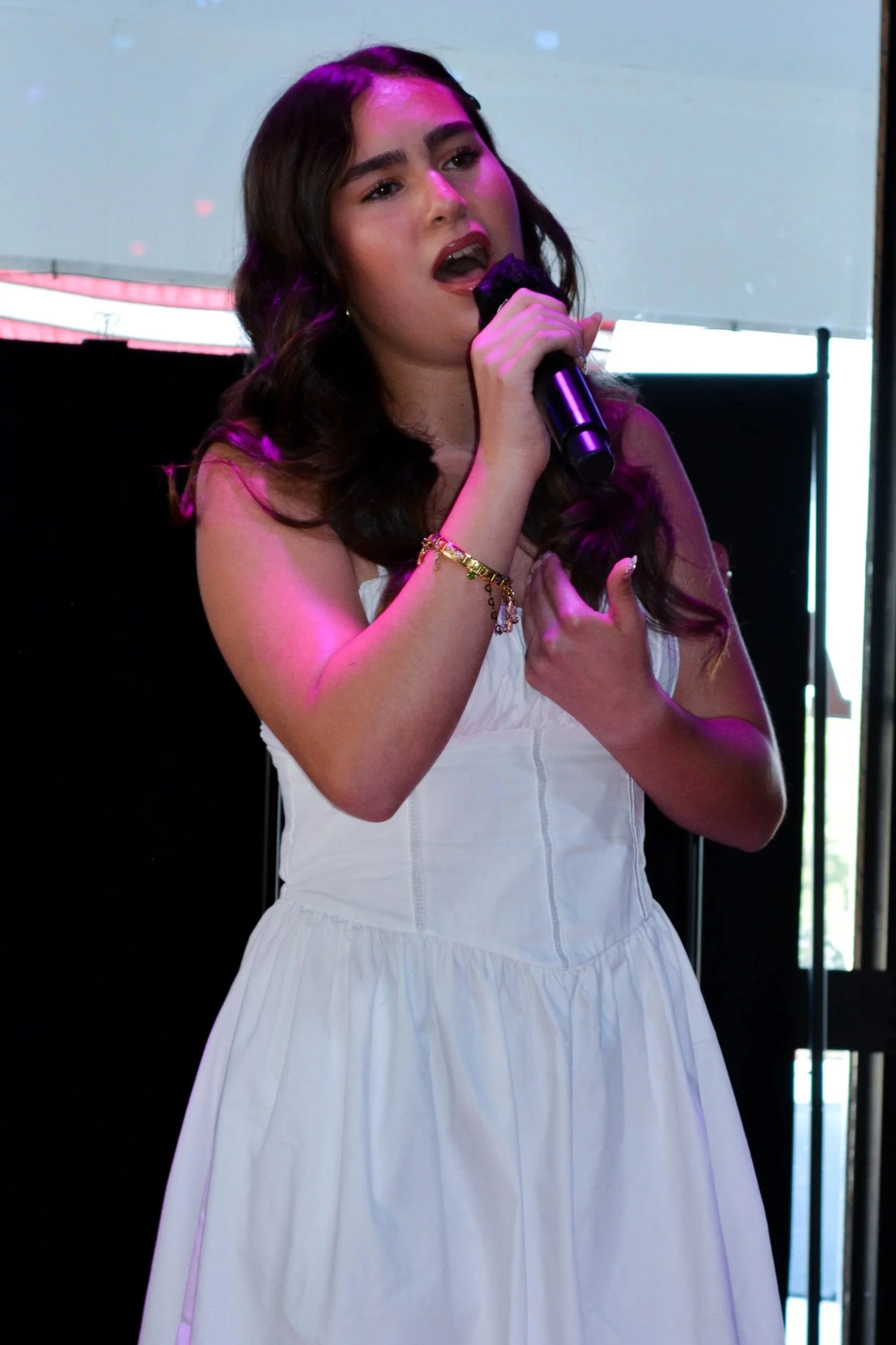 A young woman with long dark hair singing into a microphone, wearing a white dress and gold jewelry, with a background featuring dark and bright lights.