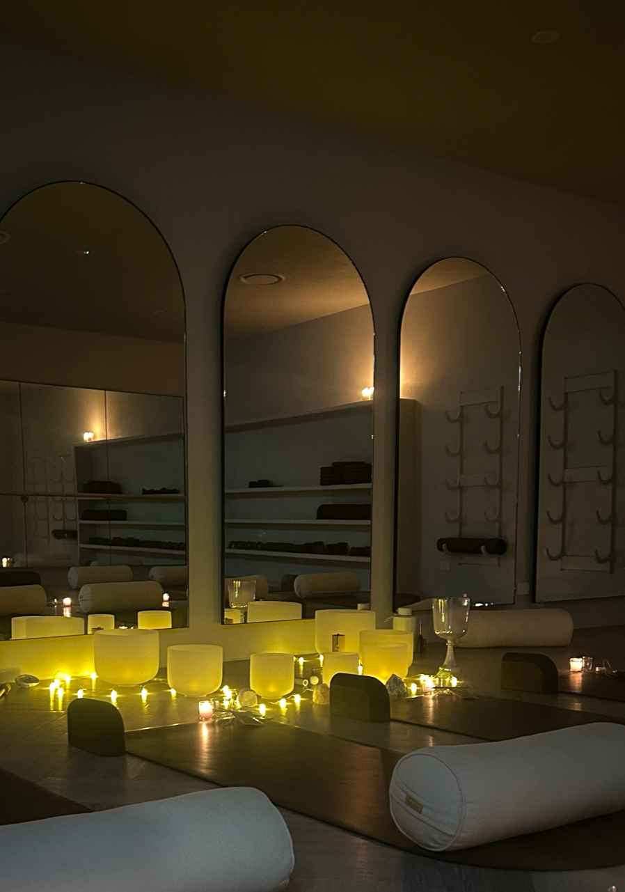 A softly lit yoga or meditation room with candles, singing bowls, and pillows on the floor, with large mirrors and minimalist decor.