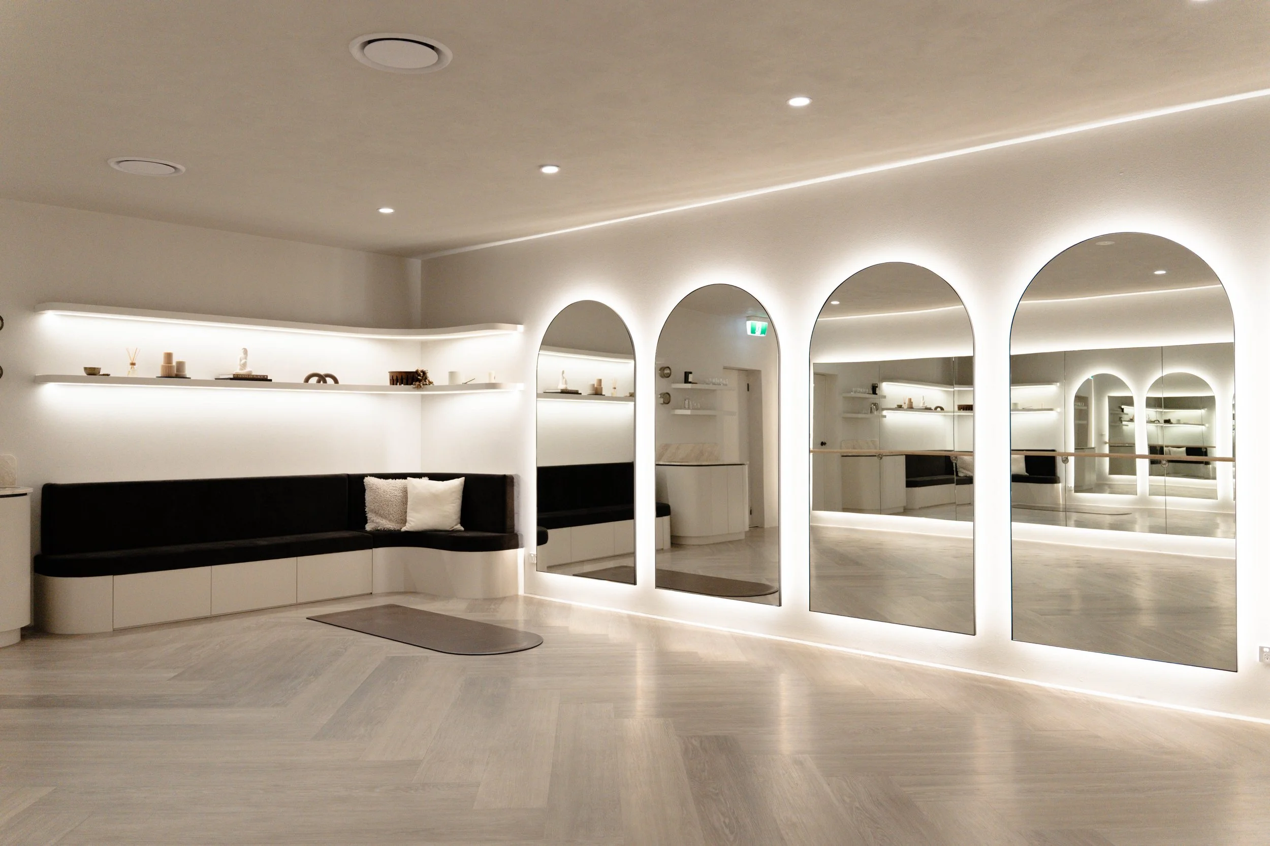A modern, minimalistic interior with white walls and ceiling, illuminated by recessed lighting. A curved black and white seating area with decorative pillows is in front of a long white shelf displaying small decorative objects. Large mirror panels with arched tops reflect the room.