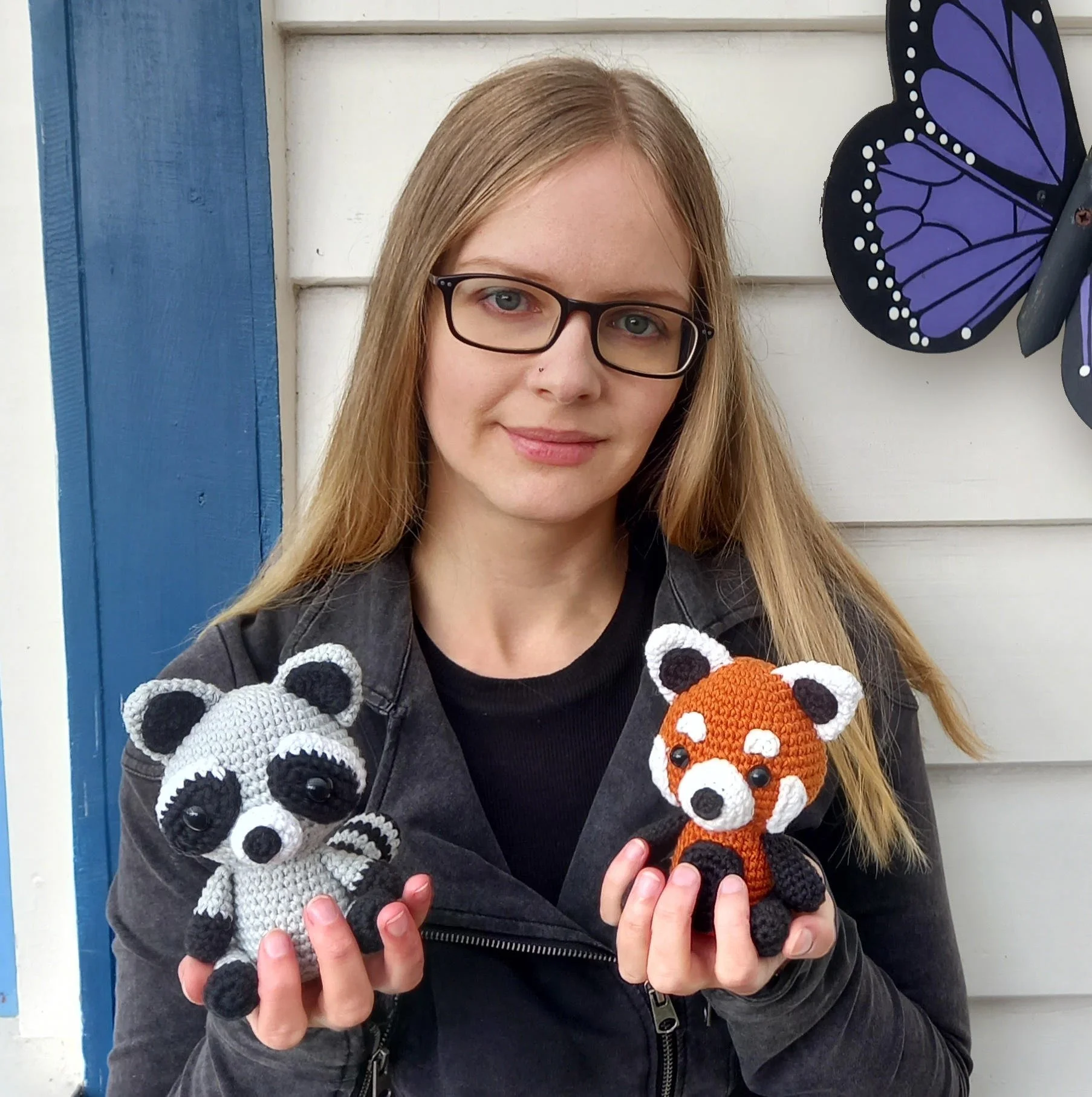 A woman wearing glasses and a black jacket holding two crochet animal plush toys, a raccoon and a fox, standing in front of a white wall with a blue trim. A butterfly decoration is partially visible on the right side.