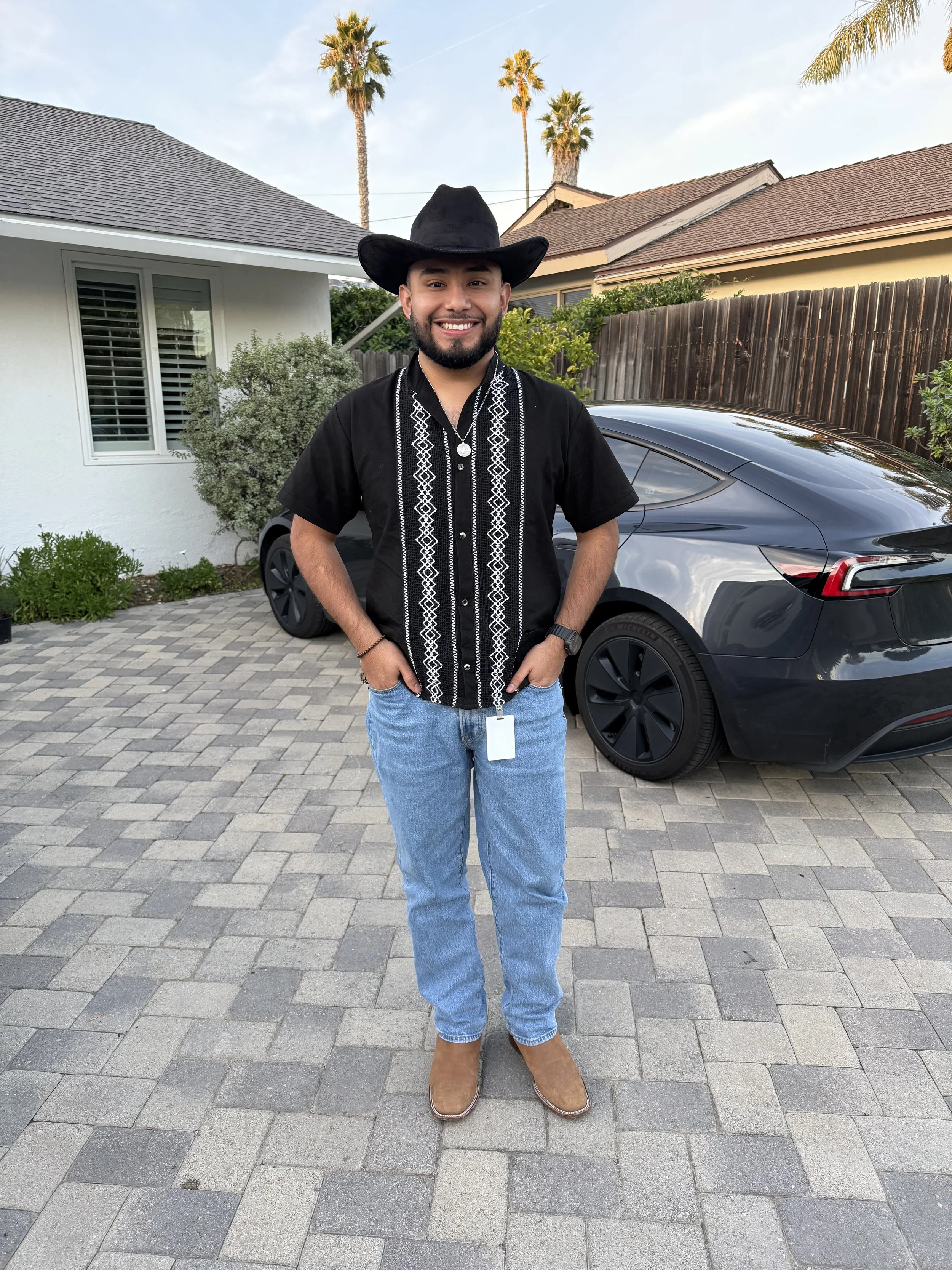 A man dressed in western attire, wearing a black cowboy hat and black shirt with white embroidery, standing on a paved driveway in front of a black car. There are houses, palm trees, and a wooden fence in the background.