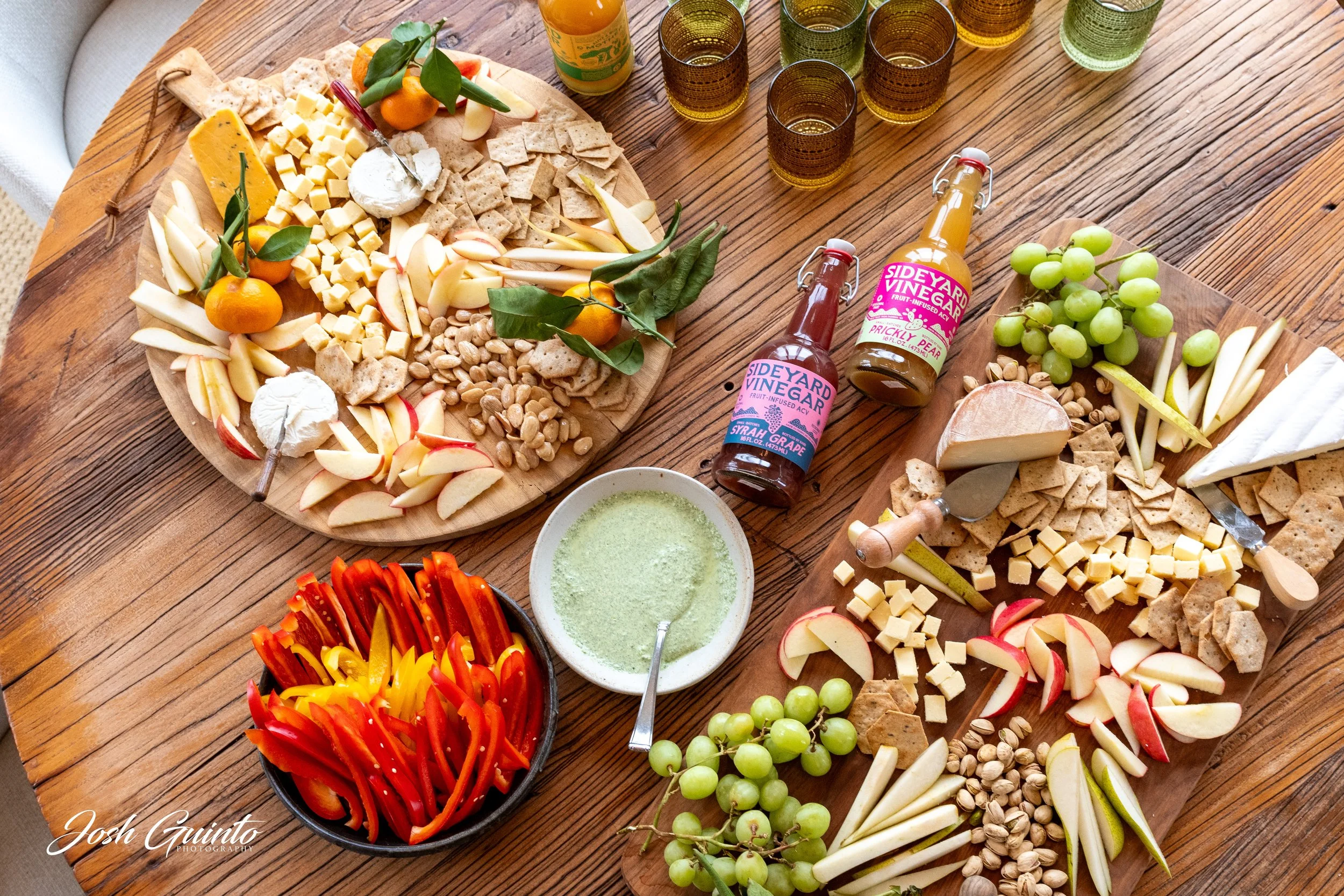 Cheese, crackers, apples, grapes, pear slices, pistachios, and honey mustard arranged on wooden serving boards for a cheese platter. There are also bottles of fruit vinegar, a bowl of green dip, and glasses of drinks on a wooden table.