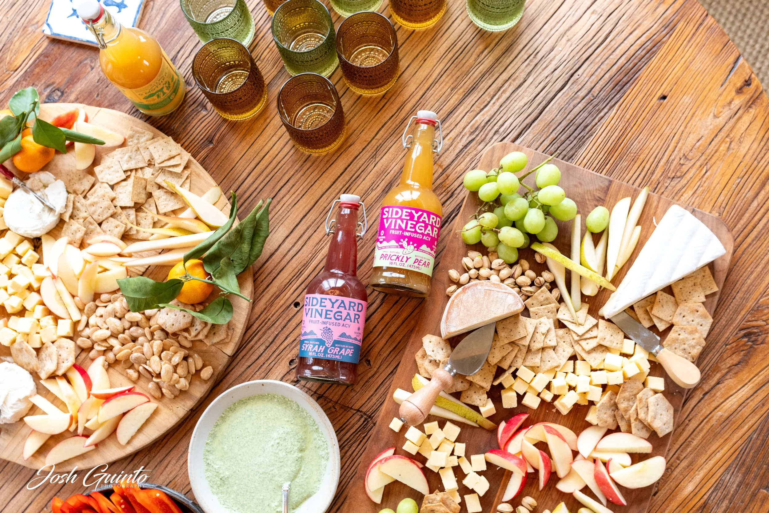 Cheese platter with sliced apples, grapes, nuts, crackers, and cheeses, with bottles of raspberry and pear flavored vinegar, and glasses of a yellow beverage on a wooden table.