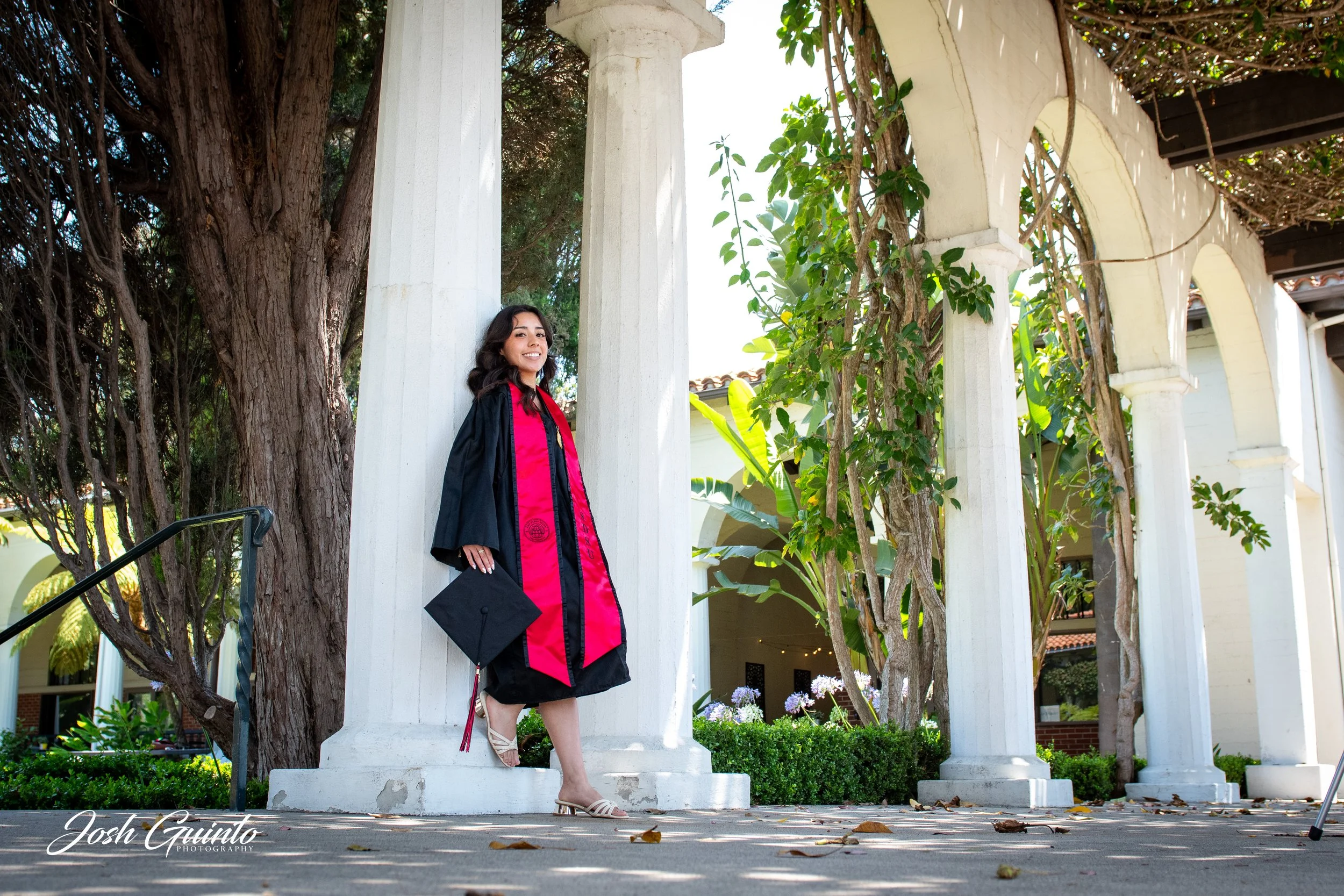 A woman in a black graduation gown and pink stole stands outside on a walkway with white columns and greenery, smiling, holding a black graduation cap.
