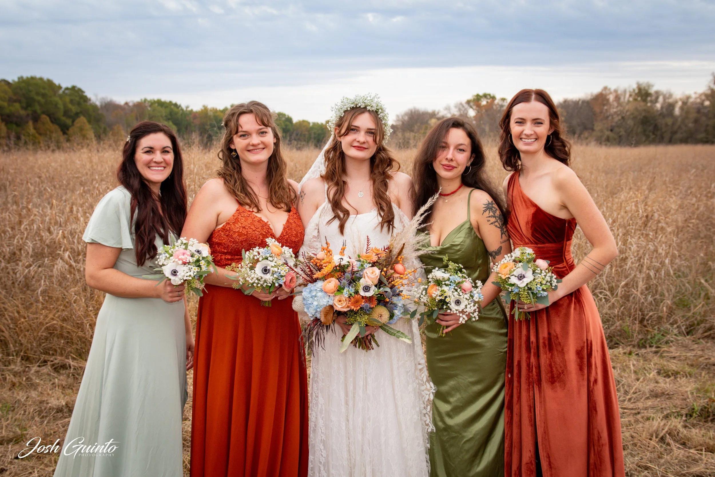 Group of five women in colorful dresses standing in a field with cloudy sky, holding bouquets, with one woman in a white dress and floral headband in the center.