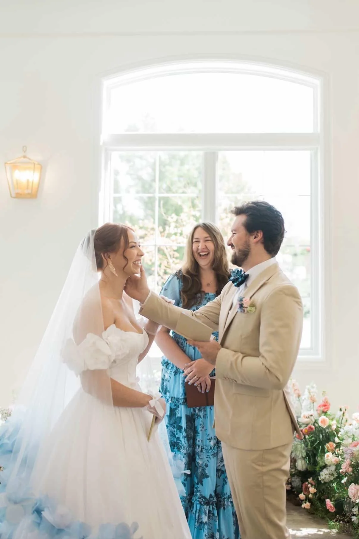 A bride and groom exchanging vows with a woman in blue officiating in front of a large window with flower arrangements nearby.