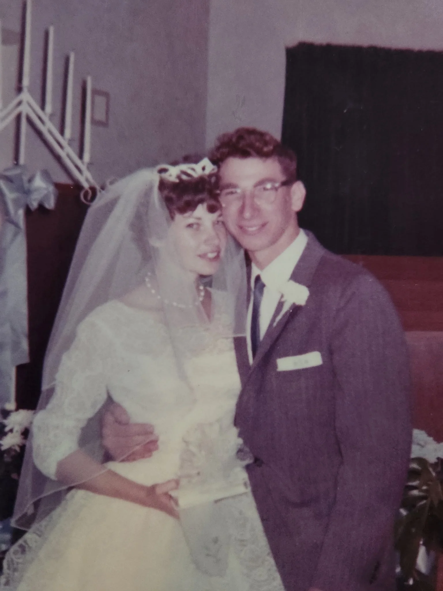 A bride and groom dressed in wedding attire, posing together indoors. The bride is wearing a white wedding dress with lace details, a veil, pearl necklace, and pearls in her hair. The groom is wearing a dark suit with a white shirt, tie, and boutonniere. They are smiling and standing close to each other.