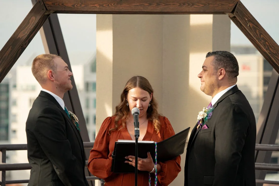A same-sex couple exchanging vows during their wedding ceremony in front of an officiant on a rooftop, with an urban cityscape in the background.