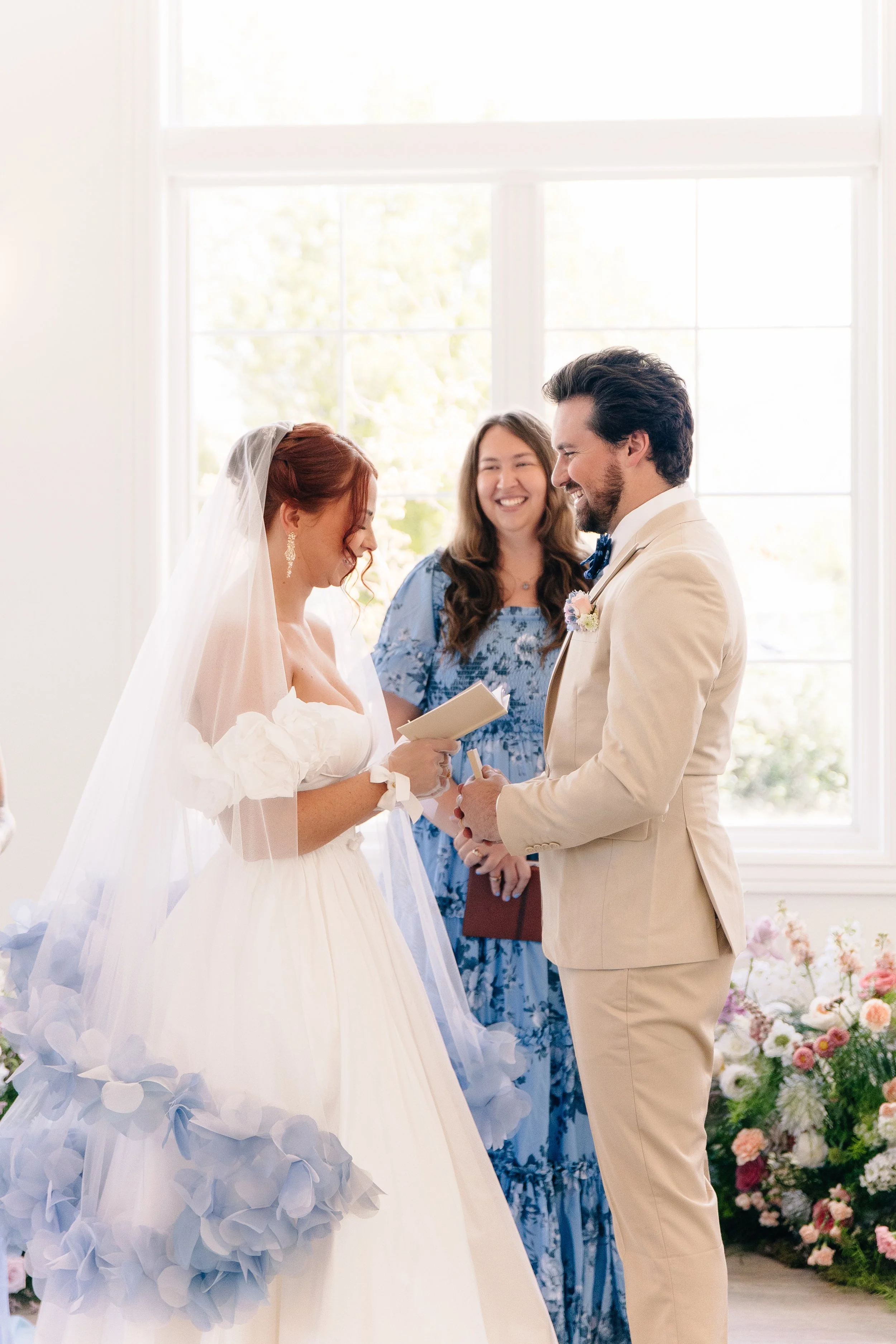 A bride and groom exchanging vows during their wedding ceremony indoors with large windows and flowers, with an officiant standing behind them.