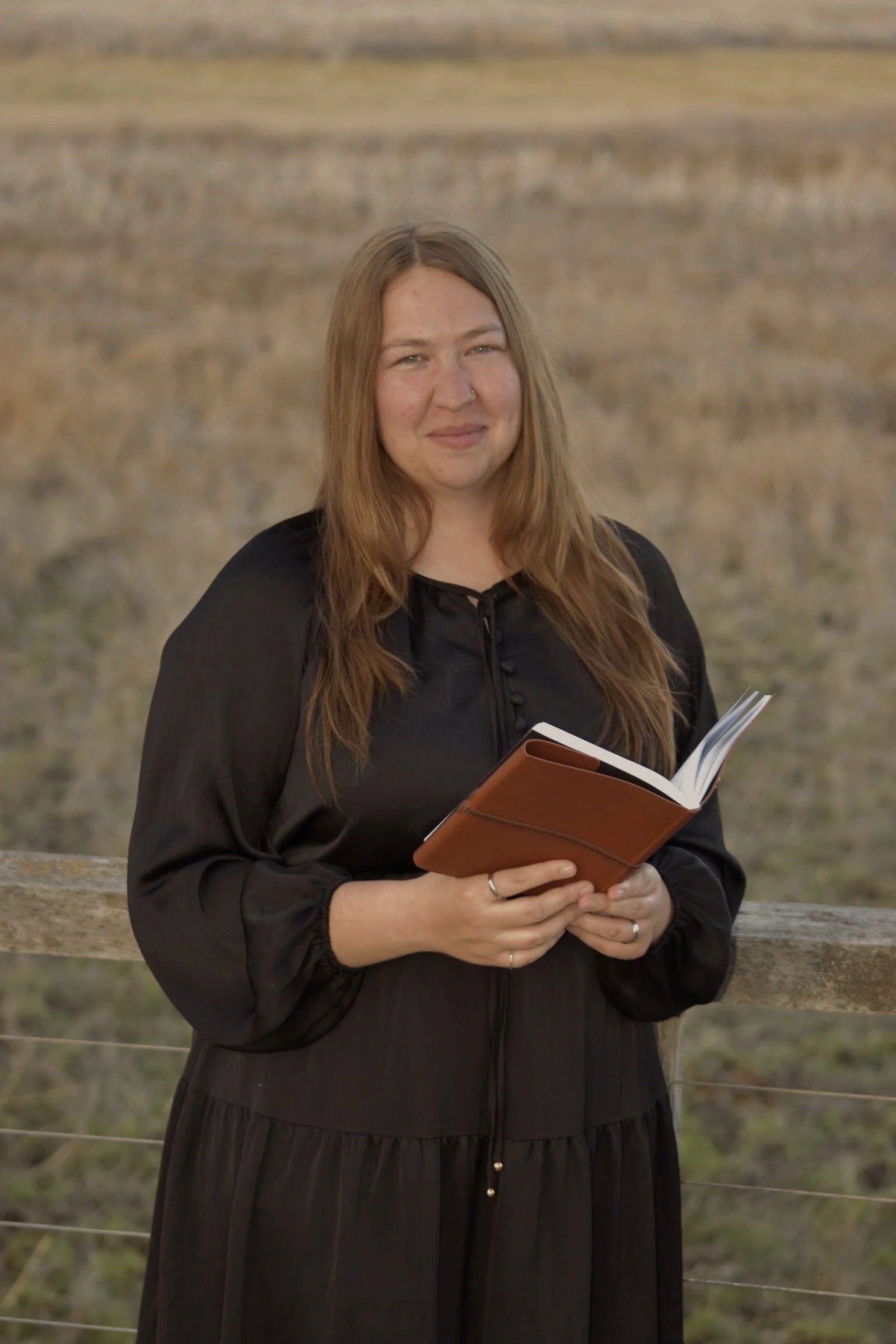 A woman with long brown hair and a nose piercing holding an open notebook and standing outdoors near a wooden fence with a field in the background.
