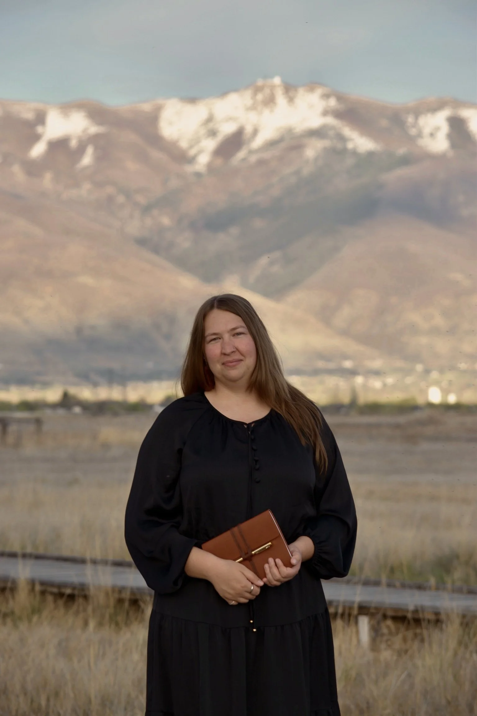 A woman with long brown hair in a black dress holding a brown notebook with a gold pen standing outdoors with mountains in the background.