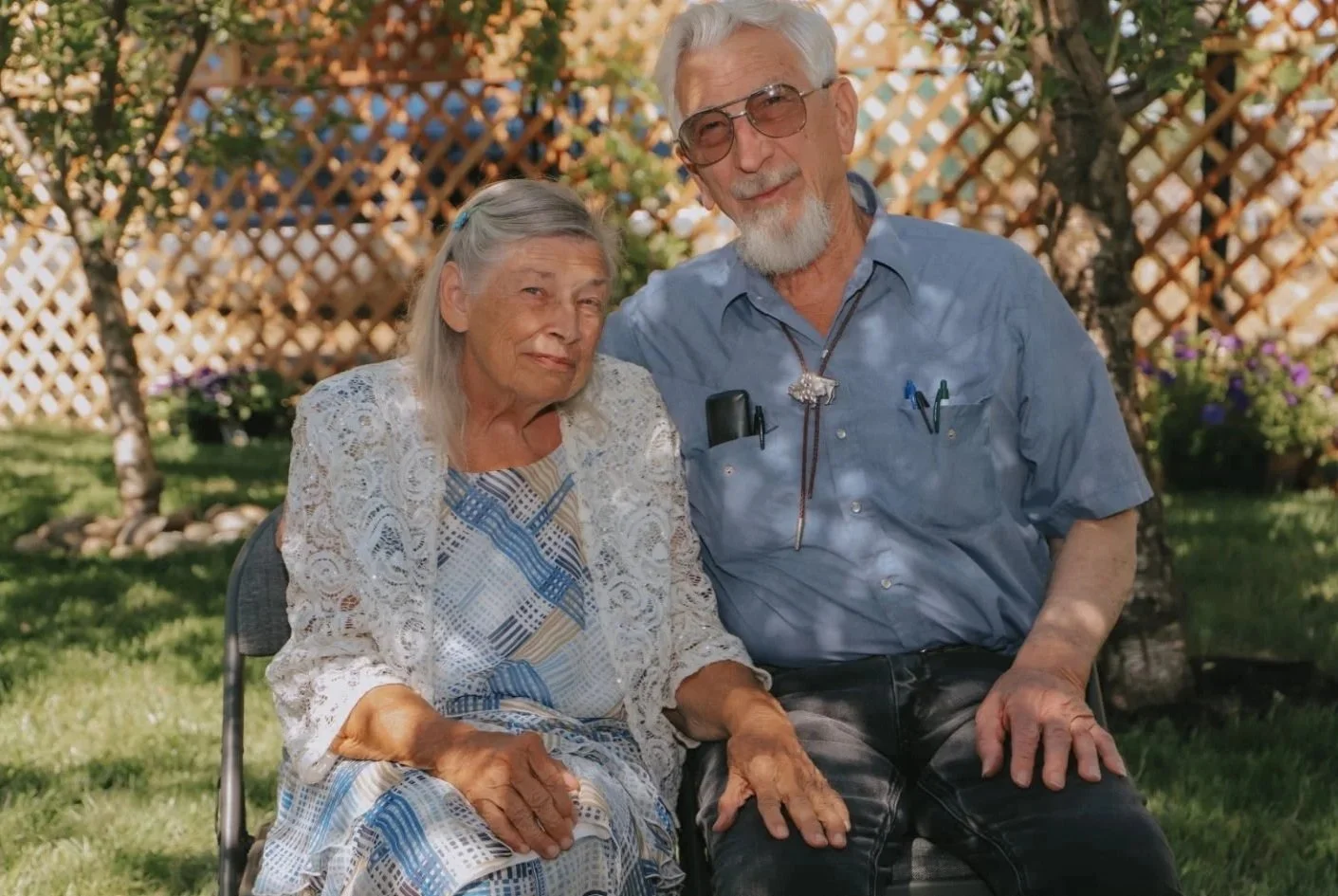 An elderly woman and an elderly man sitting outdoors on a sunny day, in front of a wooden lattice fence with trees and plants in the background. The woman is wearing a patterned dress with a white lace cardigan, and the man is wearing a blue button-up shirt with pens in the pocket, glasses, and has a white beard and hair.