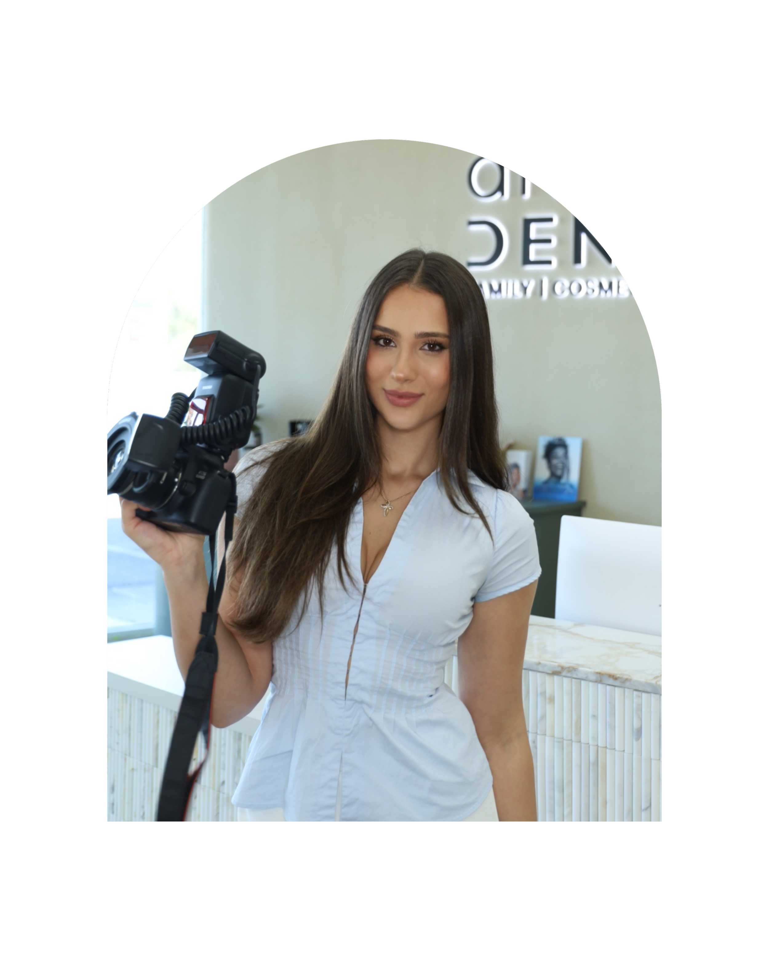 A woman with long brown hair and brown eyes wearing a white top and necklace, holding a camera, with a bright room and framed photographs in the background.