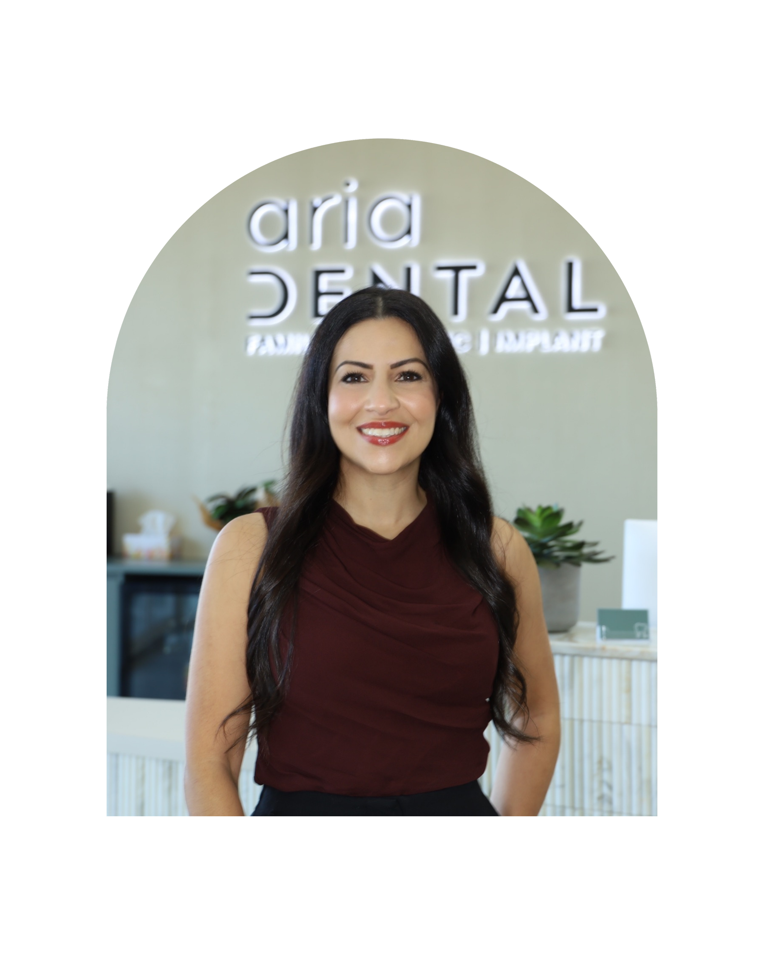 A woman with long dark hair smiling, standing in front of a sign that reads 'Aria Dental' in a dental office.