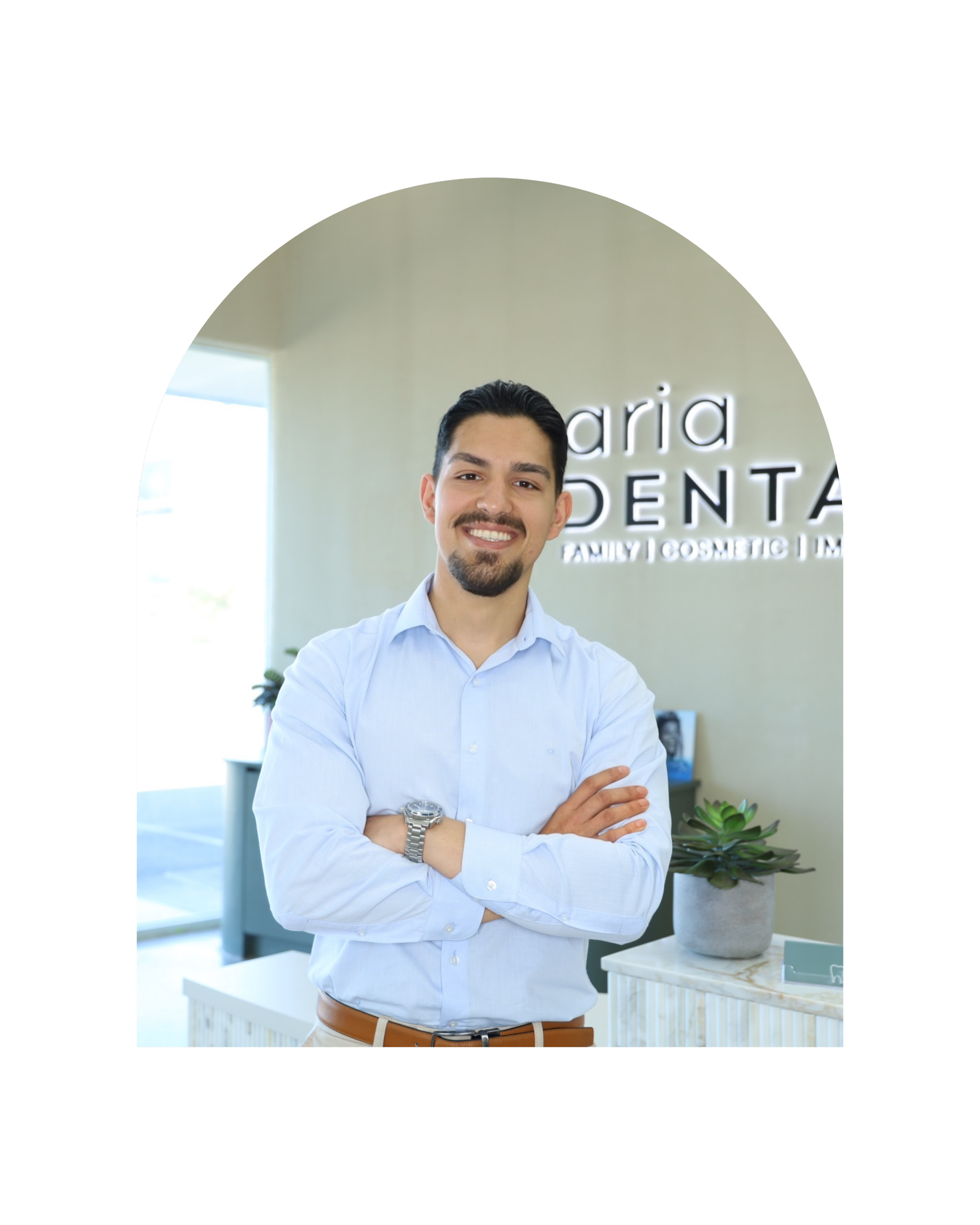 Smiling man in light blue dress shirt with crossed arms standing in an interior space with a counter and a potted succulent plant, with the word "Aria Dental" illuminated in the background.