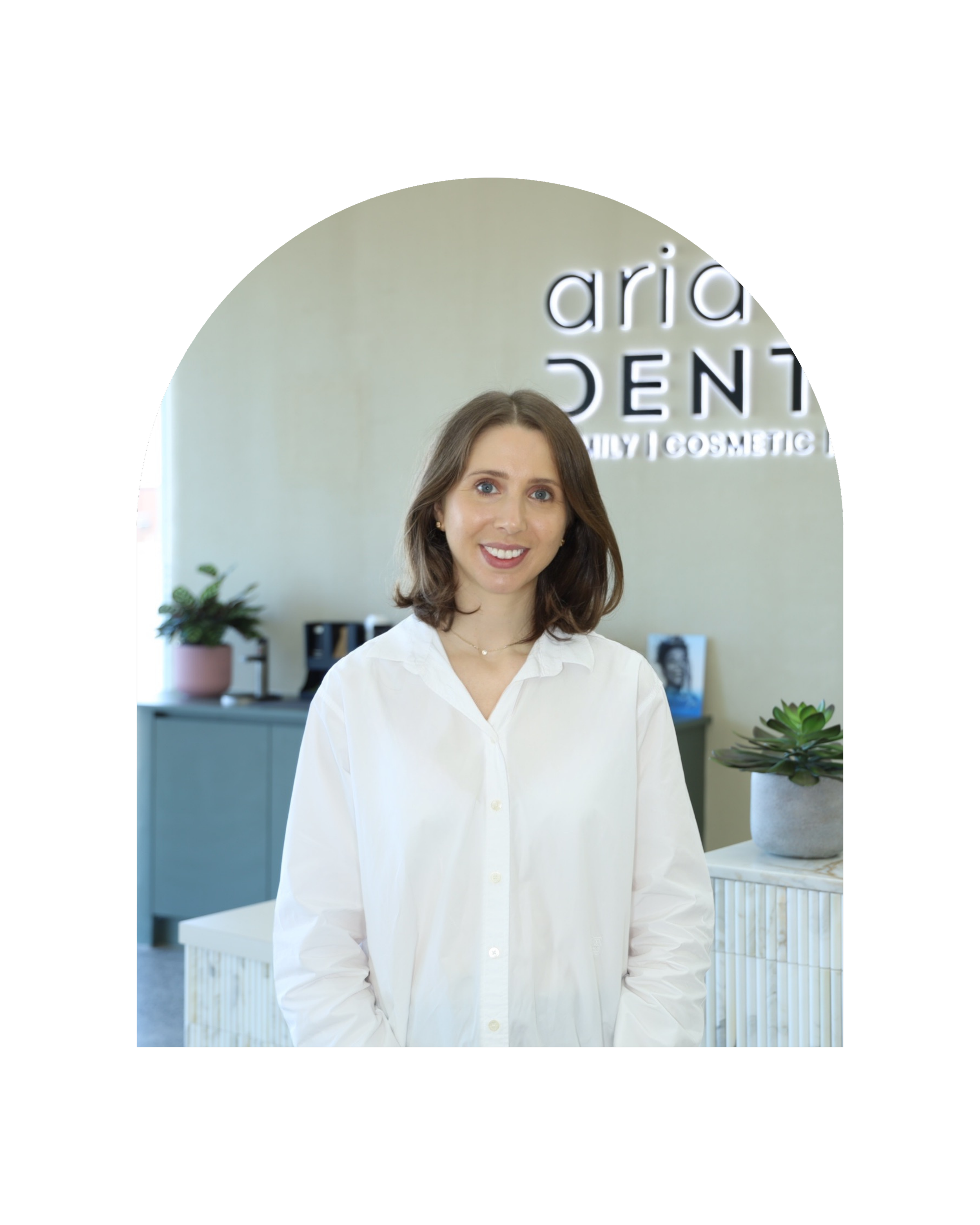 A young woman with shoulder length brown hair and blue eyes smiling, wearing a white shirt, standing indoors in front of a wall with the words "Aria Dental" illuminated behind her. There is a potted plant on a side table to her right.
