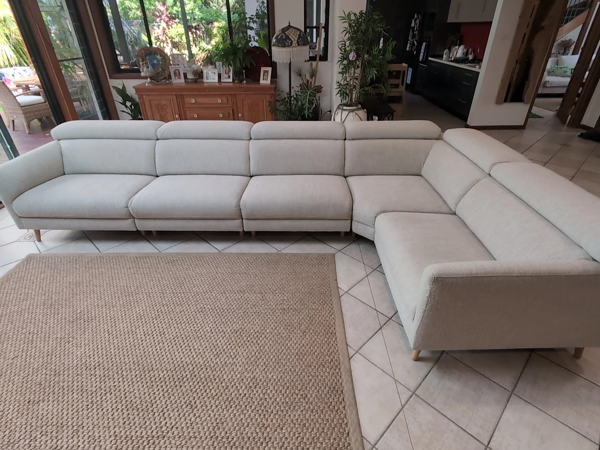 Living room with white sectional sofa, beige rug, wooden sideboard with family photos, green plants, and large window with patio view.