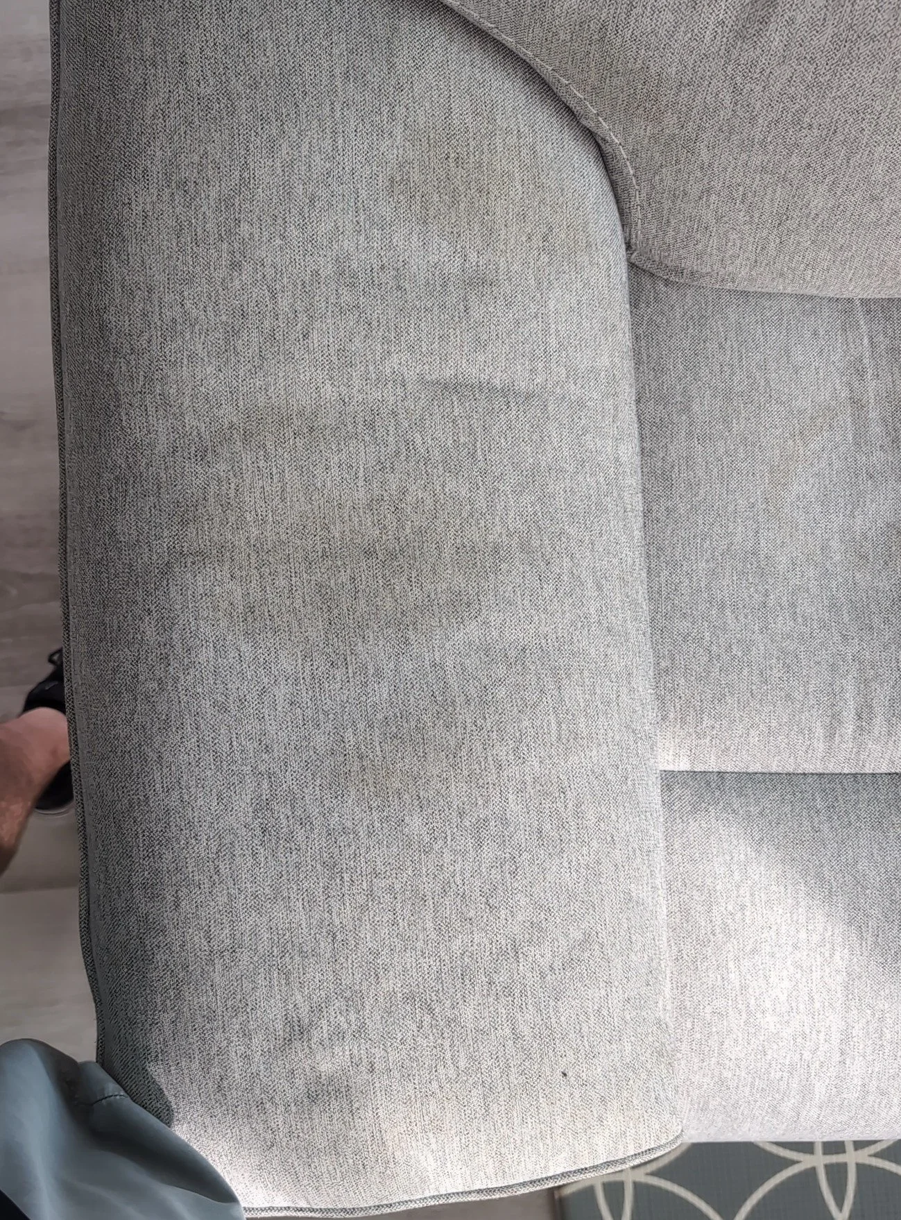Close-up of a gray fabric armrest of a sofa with a patterned carpeted floor visible at the bottom.