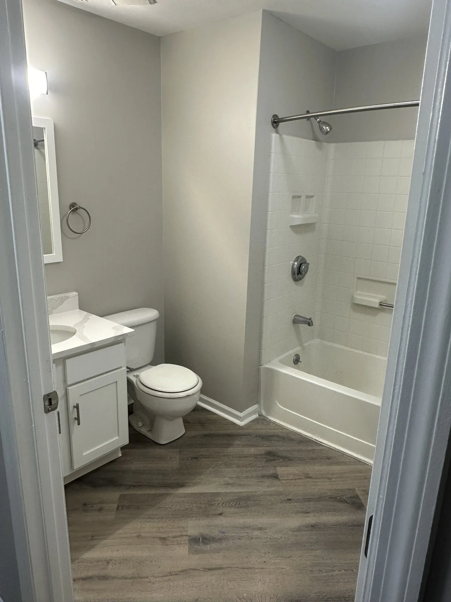 A small bathroom with a toilet, a white vanity with a sink, a mirror, a towel ring, and a bathtub with a shower, all in neutral tones and wood-like flooring.