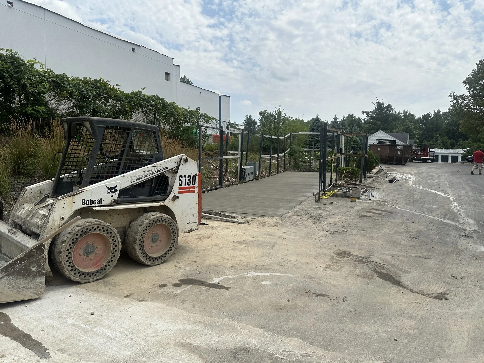 Construction site showing a Bobcat mini skid steer parked near a newly laid concrete ramp with safety fencing, adjacent to a gravel lot under a partly cloudy sky.