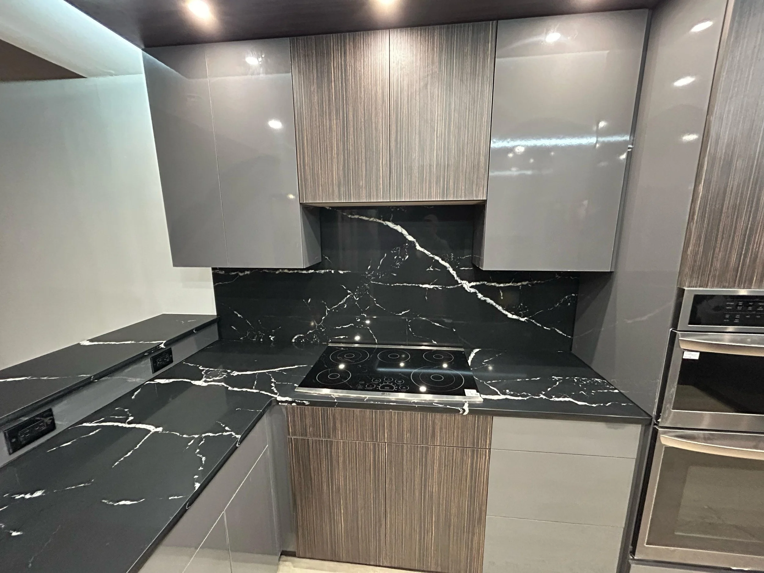 Modern kitchen with black marble countertops and backsplash, gray upper cabinets, wood-textured lower cabinets, and stainless steel appliances.