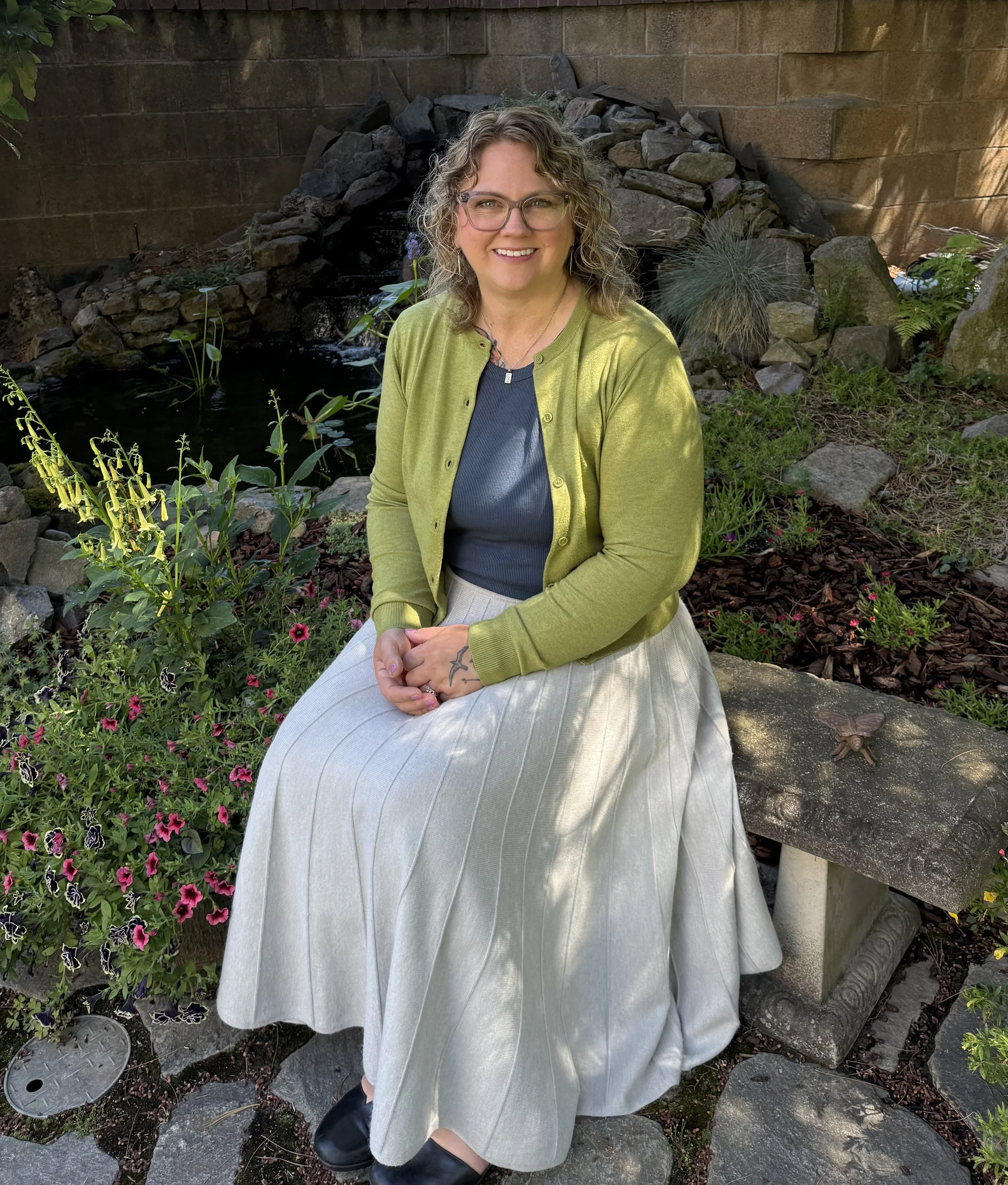 A woman with curly hair and glasses sitting on a stone ledge in a garden, wearing a light gray pleated skirt, a blue top, and a lime green cardigan, surrounded by colorful flowers and plants.