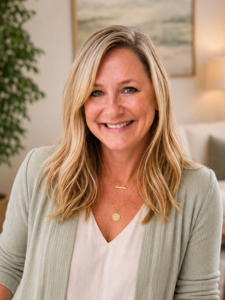 A smiling woman with blonde wavy hair, wearing a light green cardigan and layered gold necklaces, sitting in a cozy, well-lit room with a framed painting, a lamp, and a potted plant in the background.