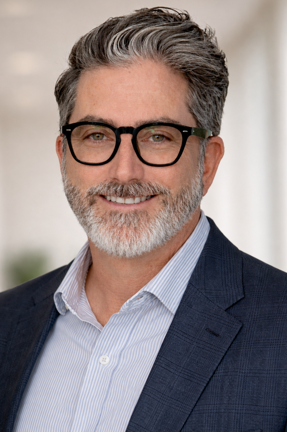 Headshot of a middle-aged man with gray hair and beard, wearing glasses, a blue blazer, and a light blue striped button-up shirt, smiling.