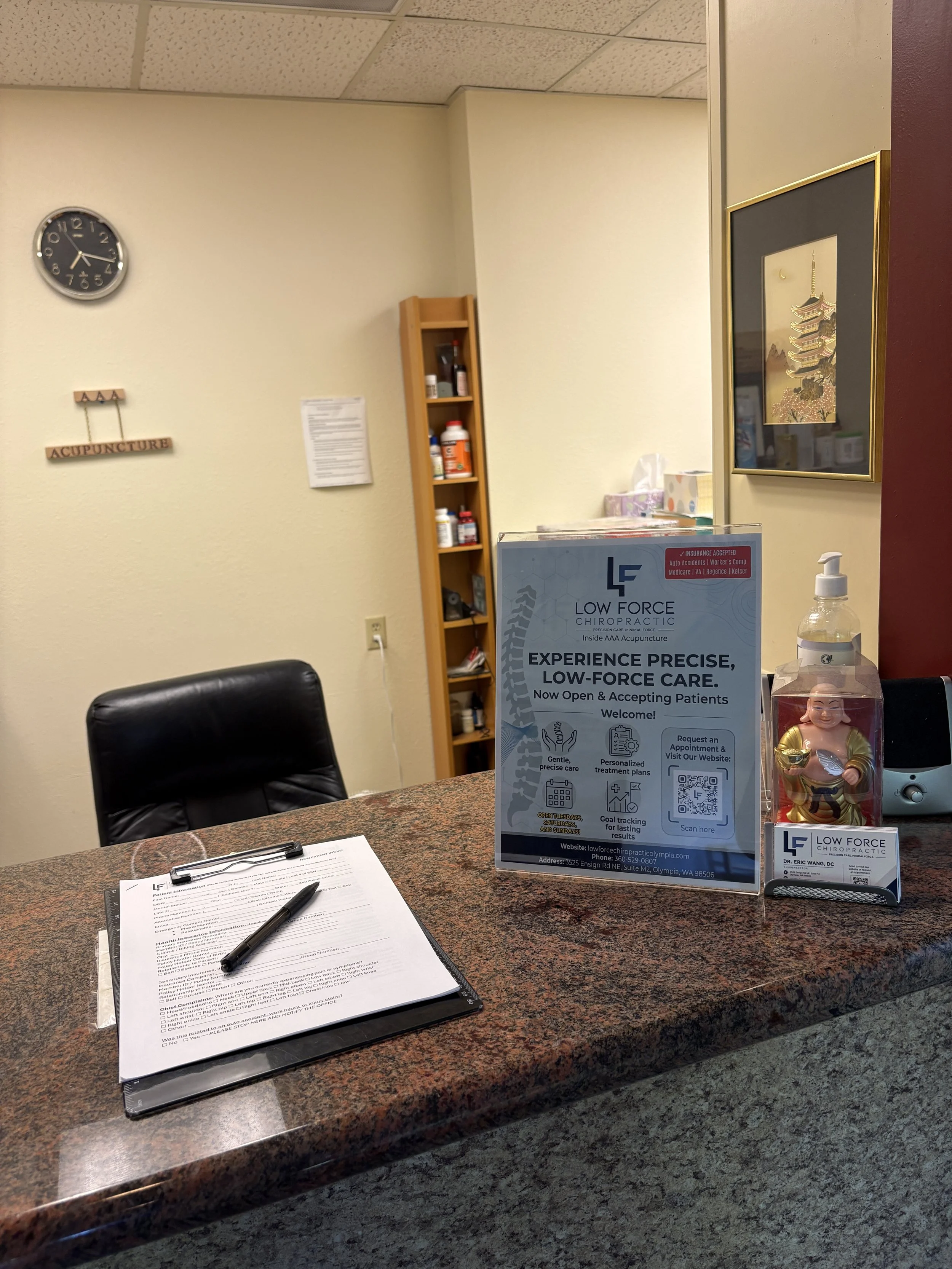Reception area of Low Force Chiropractic with a counter, informational signs, chairs, a tissue box, and decorative art on the walls.