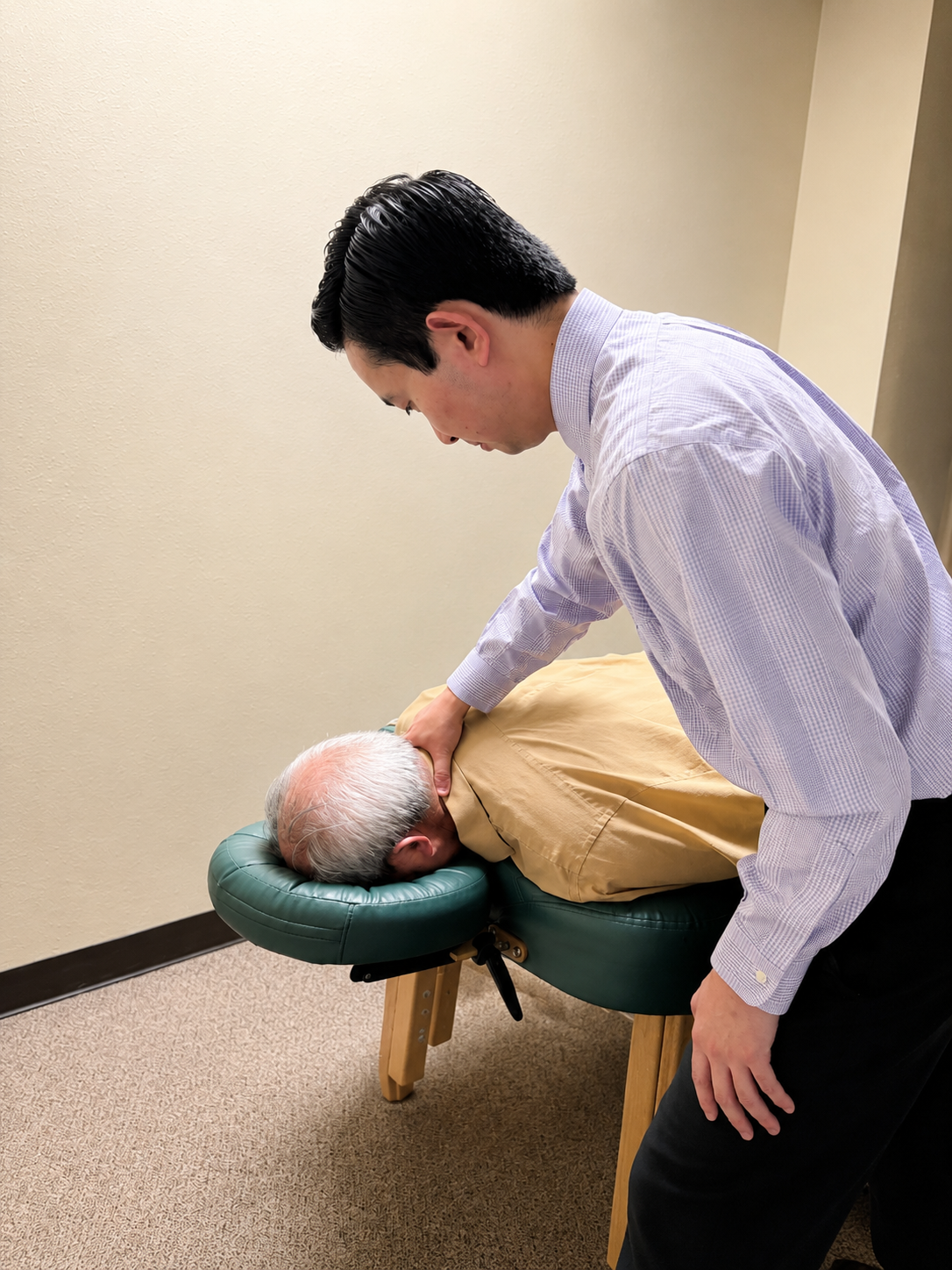 A man giving a neck massage to an elderly man lying face down on a massage table in a room with plain beige walls.