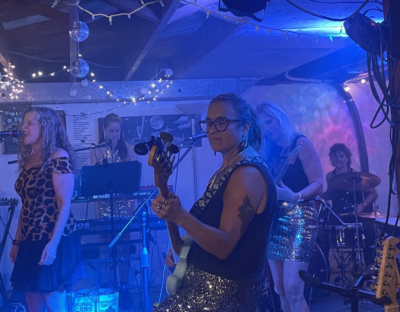 A group of women performing live music on stage in a dimly lit setting decorated with string lights and disco balls, with a woman in the foreground holding a guitar.