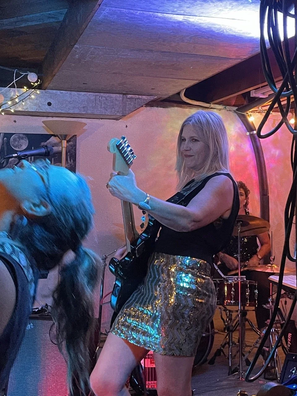 A woman with blonde hair playing an electric guitar on stage with a drummer in the background.