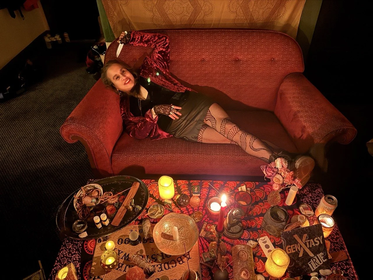 A woman lying on a red vintage sofa surrounded by lit candles, flowers, and various objects in a dimly lit room with warm lighting.