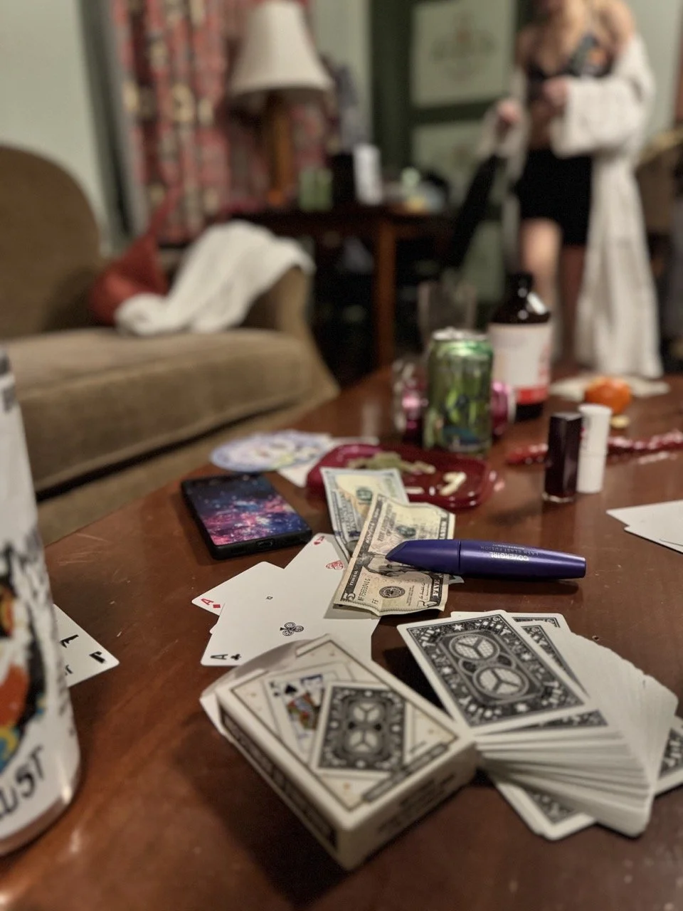 A cluttered table with playing cards, cash, a pen, and drink cans, with two blurred people in the background.