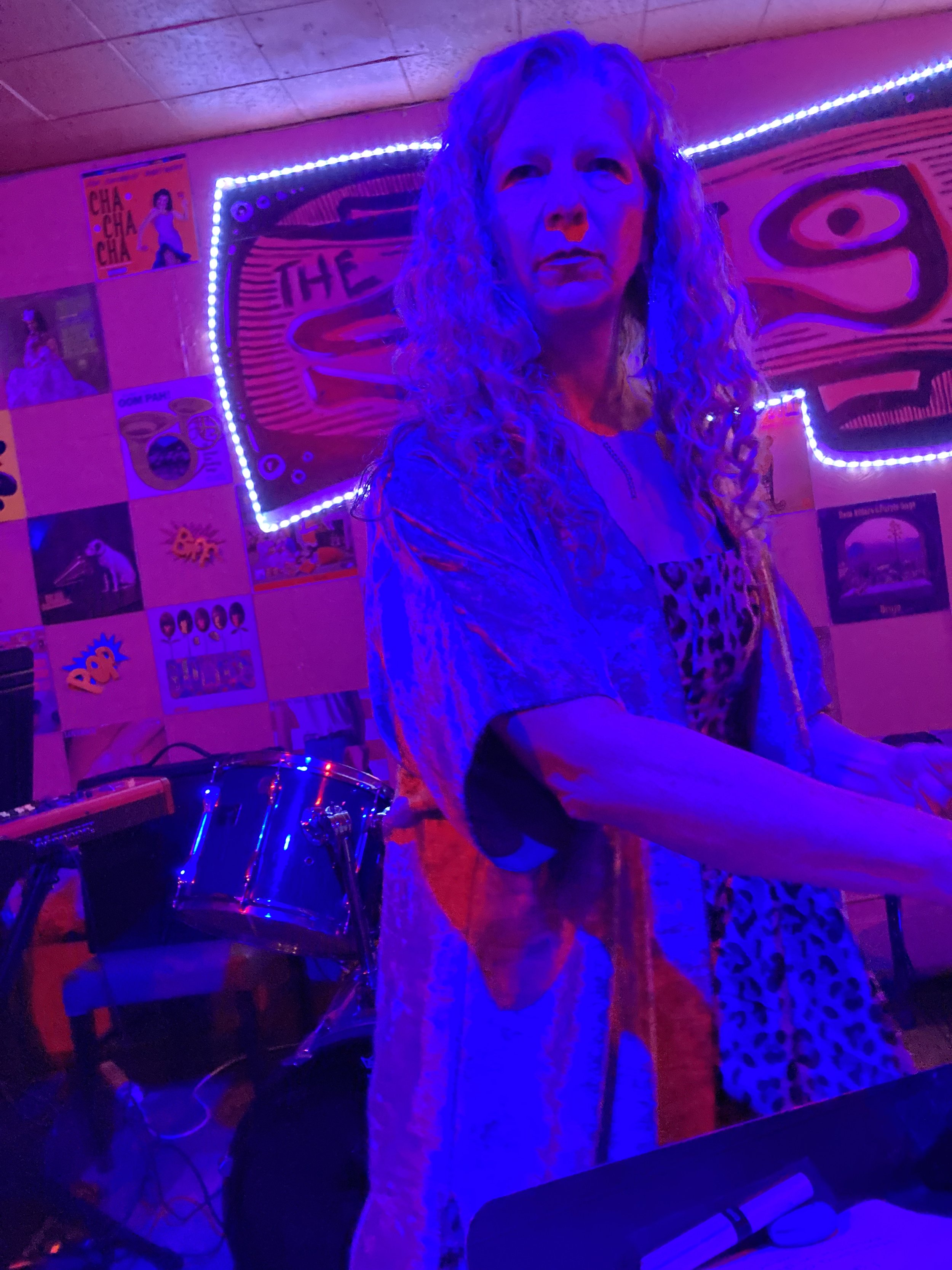 A woman with curly hair in a dimly lit room with purple and pink lighting, standing near musical instruments, with colorful posters and a neon sign on the walls.