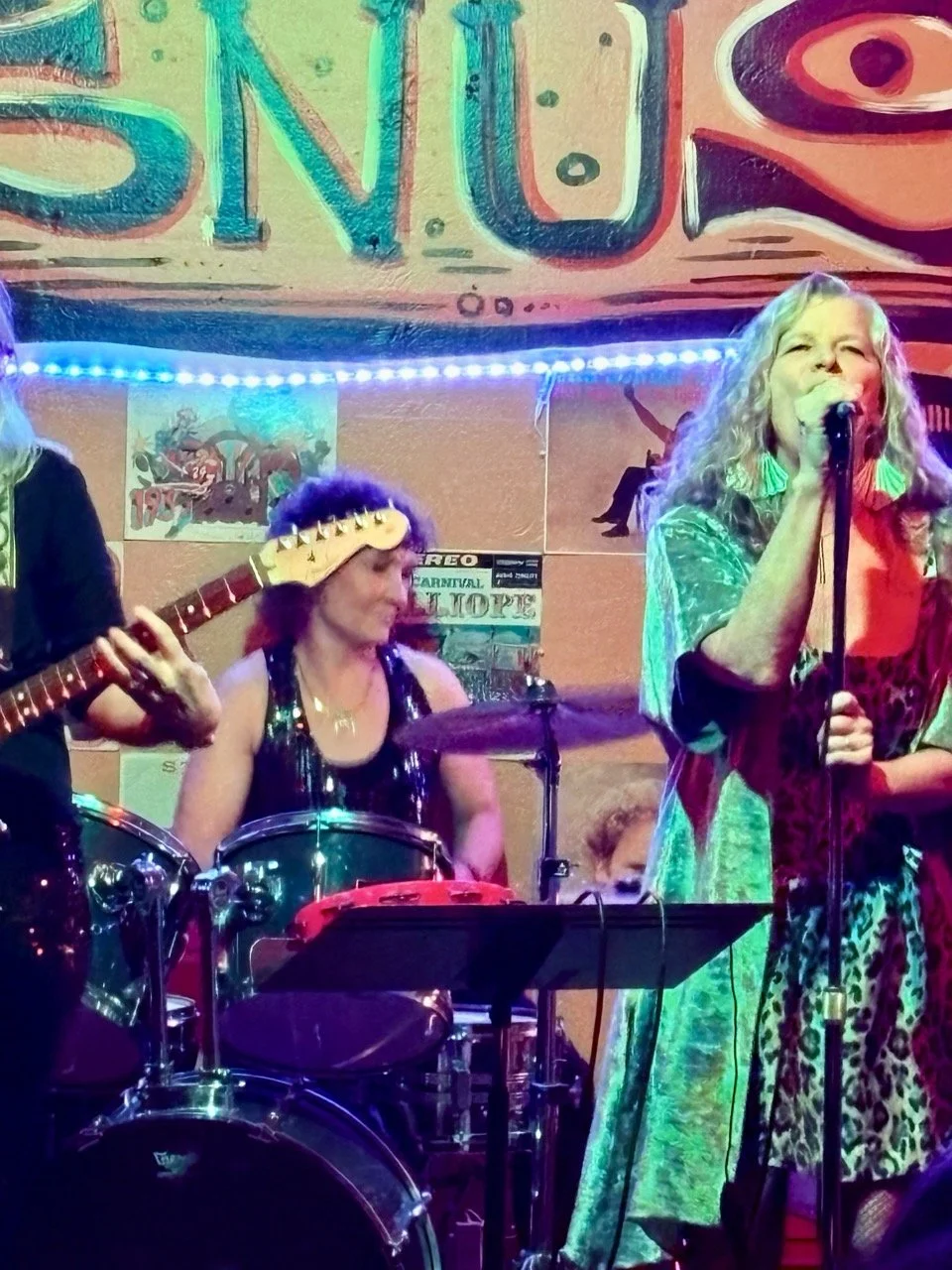 A woman with long curly blonde hair singing into a microphone on stage, wearing green earrings and a colorful leopard-print outfit. A woman with short curly hair playing drums and another person playing guitar are partially visible in the background.