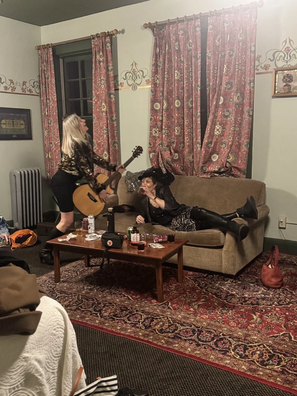 Two women in a living room, one playing guitar and the other lounging on the sofa, amid drink cans and scattered items.